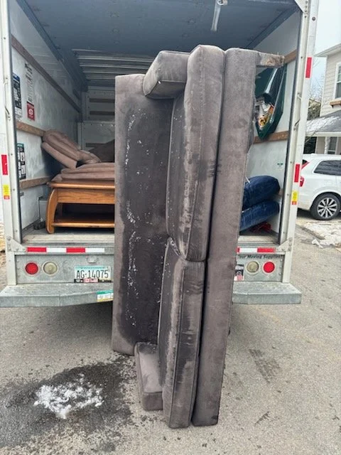 A moving truck with a large sofa on the back, leaning upright between the truck's side and the ground, with other furniture inside the truck, including a bed and a dark blue chair.