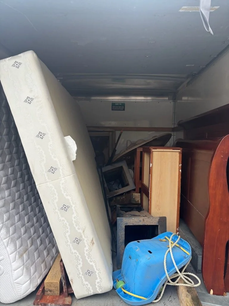 Heap of old furniture and items on a truck bed, including a mattress, a blue plastic cooler, and various wooden furniture pieces.