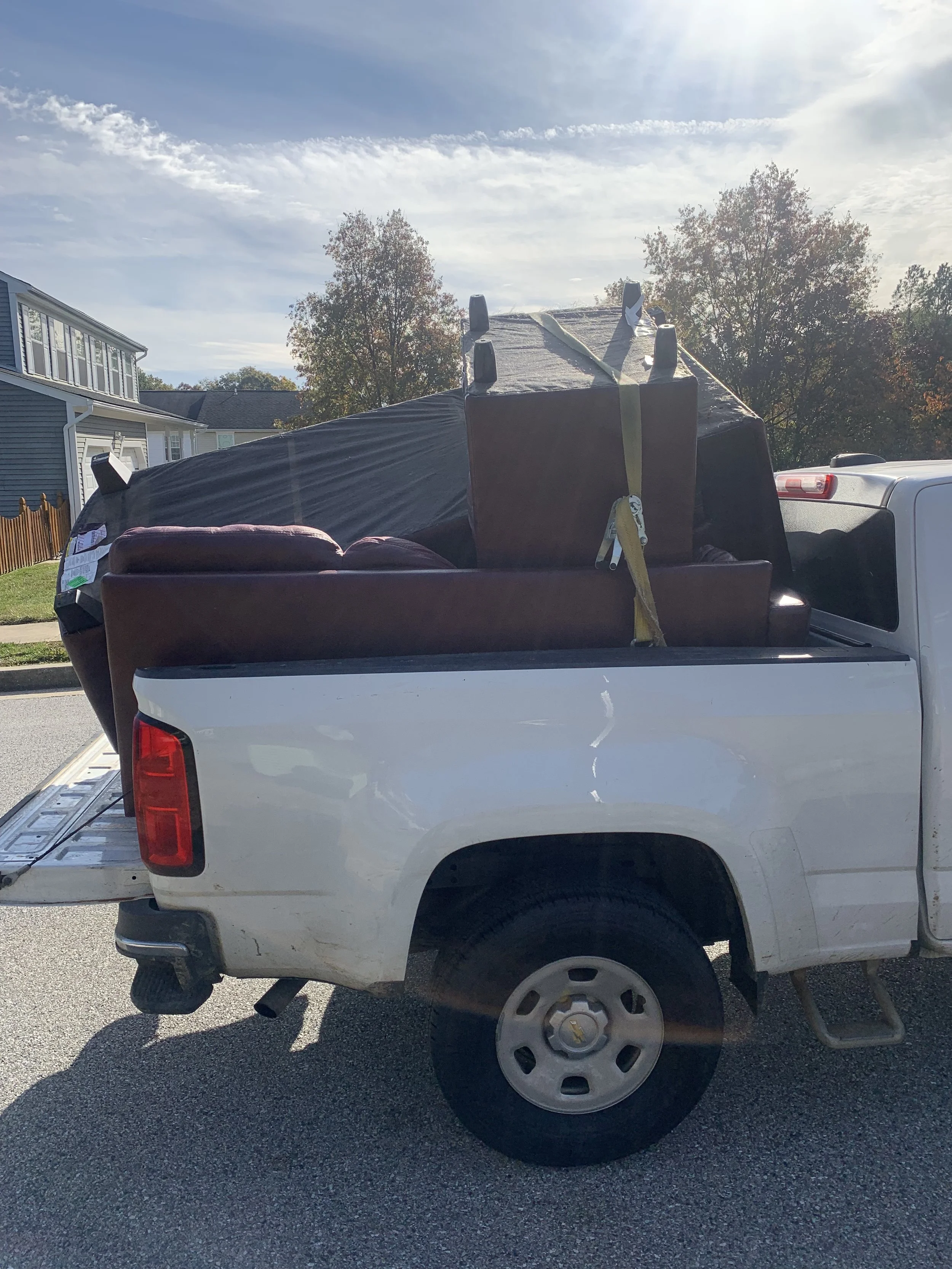 White pickup truck carrying furniture including a reclining chair and a black dresser loaded in the truck bed.