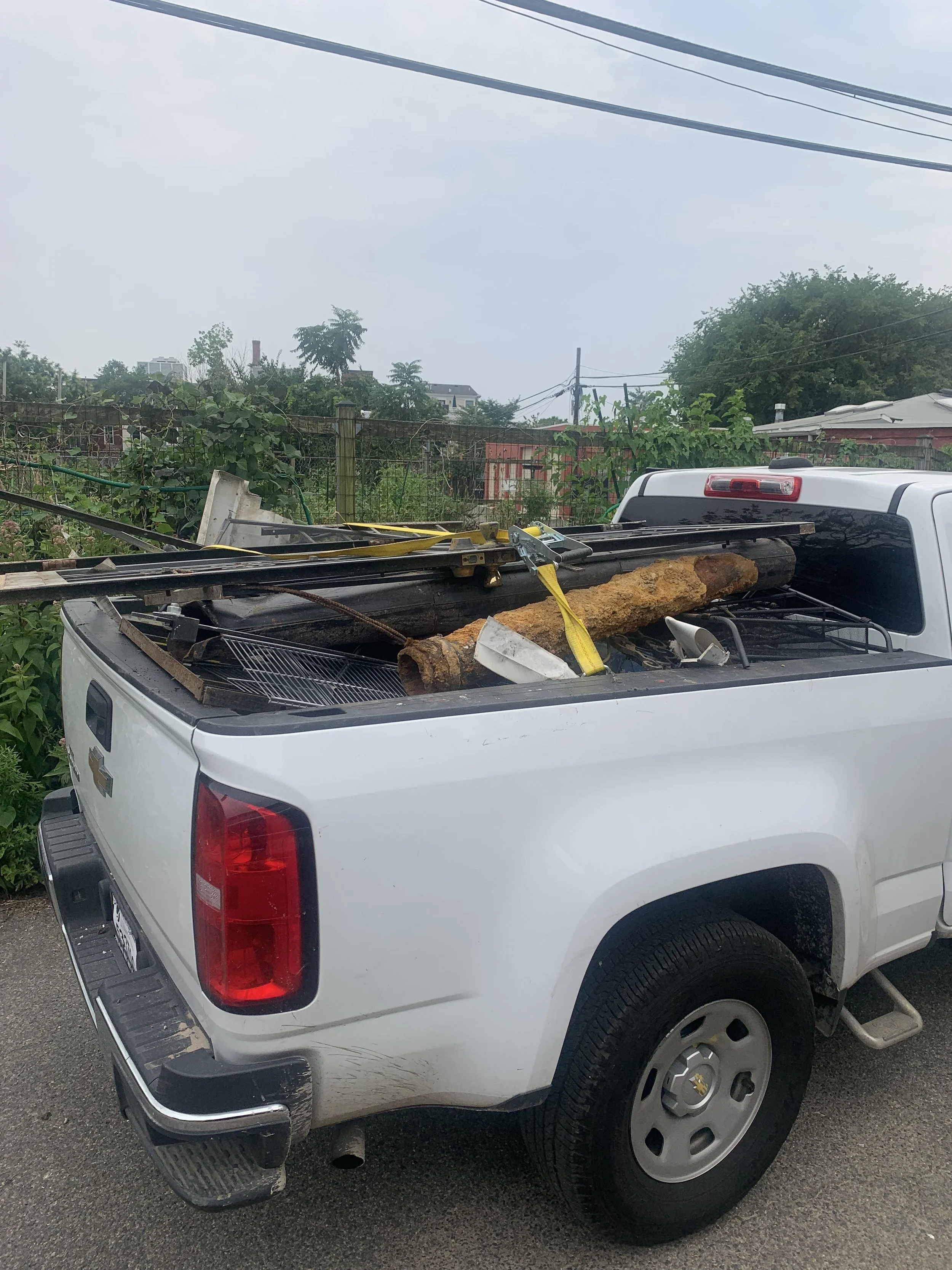 White pickup truck with cluttered cargo bed, including metal bars, a large rusty pipe, and various tools, parked outdoors with trees and power lines in the background.