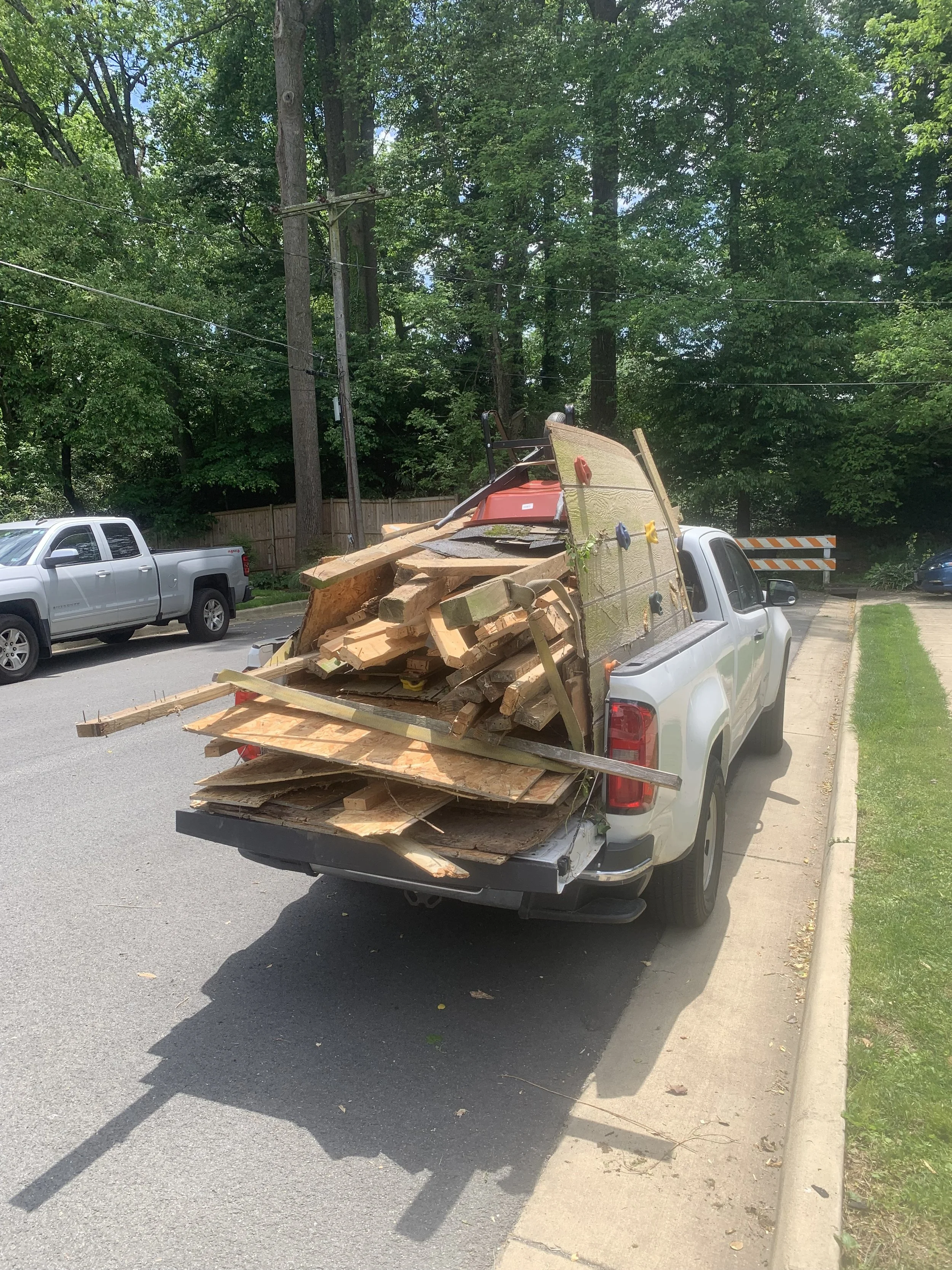 Pickup truck with loaded wooden planks, boards, and construction materials parked on a residential street near trees and other vehicles.