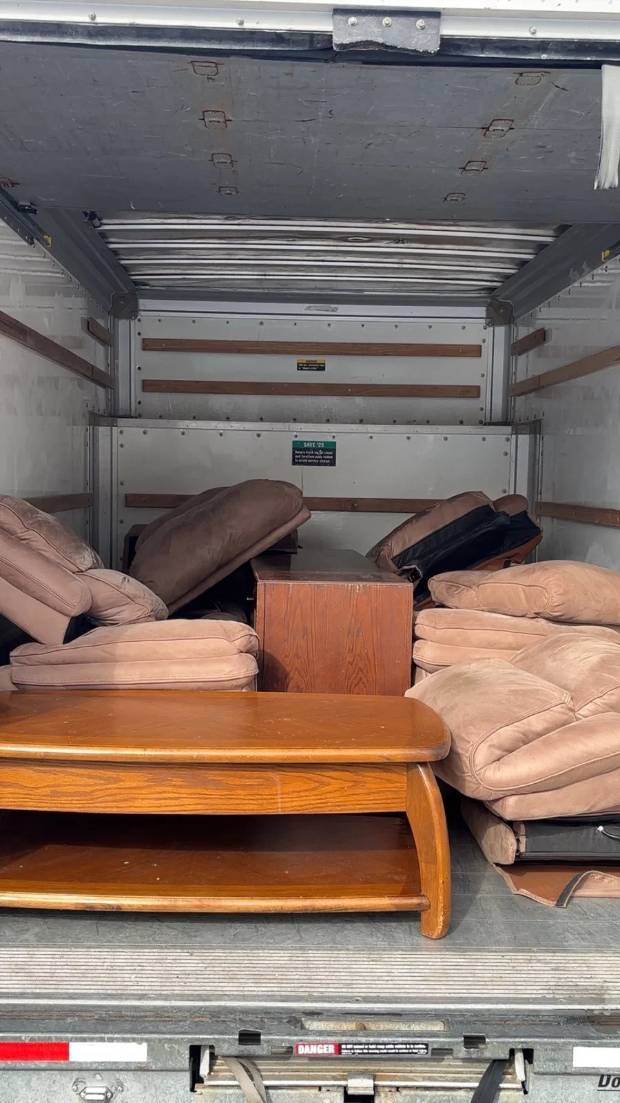 Interior of a moving truck filled with miscellaneous furniture and cushions, including wooden tables and various plush armchairs stacked and scattered.