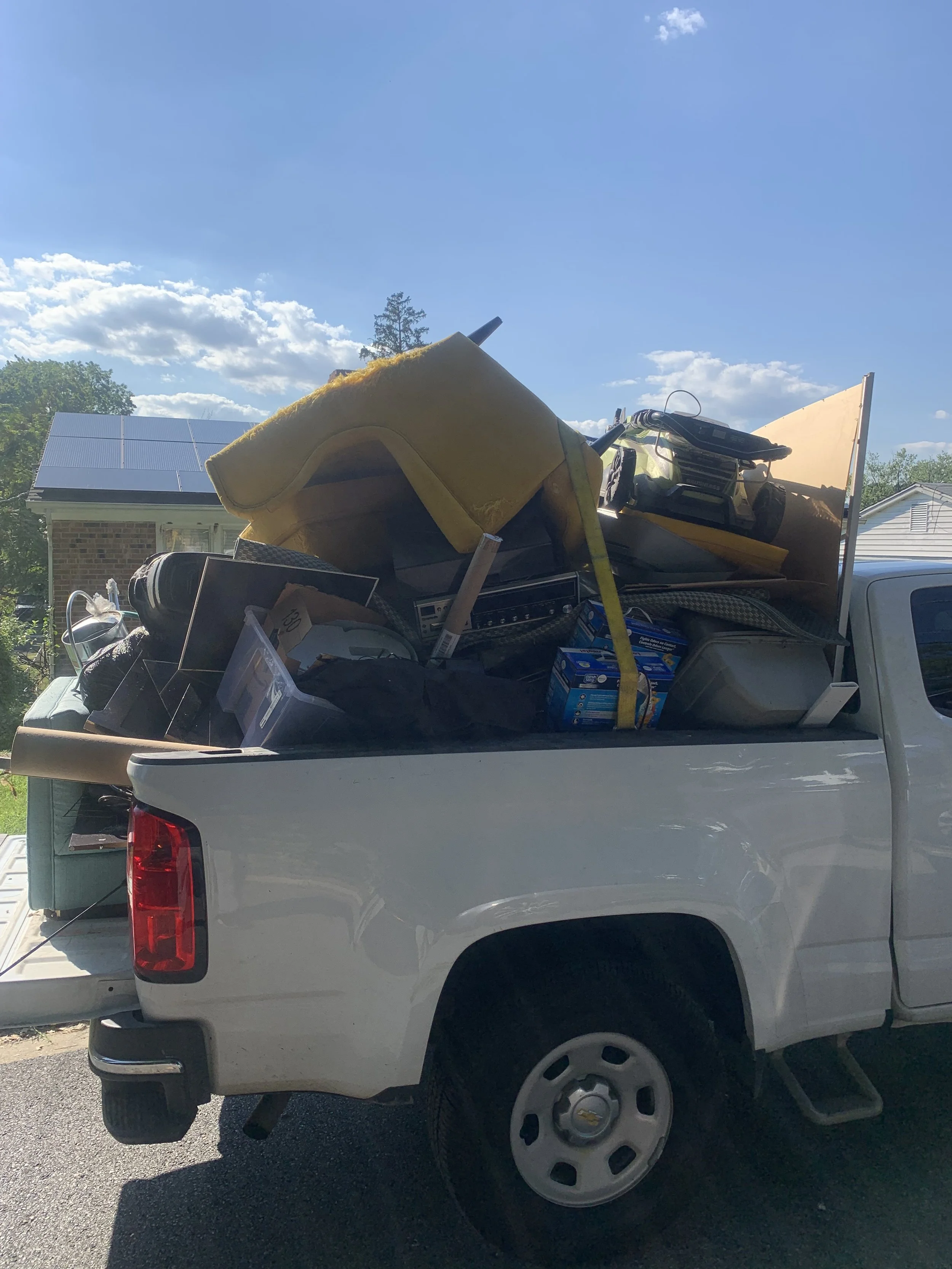 White pickup truck loaded with various household items and boxes, parked outside on a sunny day with a house and trees in the background.