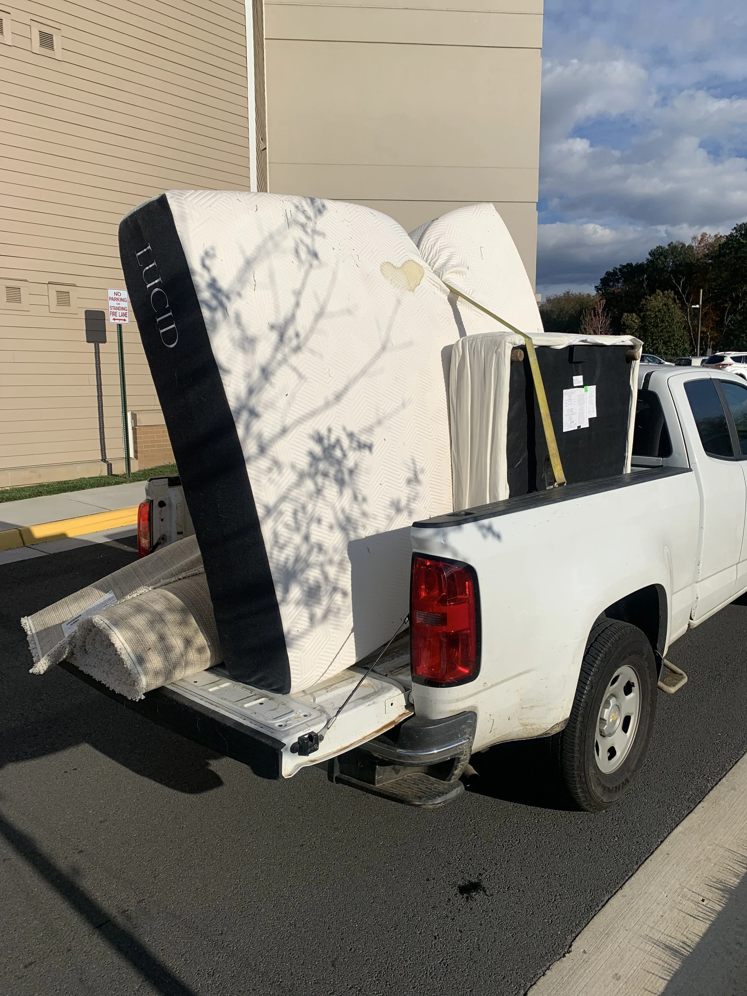 A white pickup truck parked on the street carrying a large mattress, a rolled-up carpet, and a box spring in the truck bed.