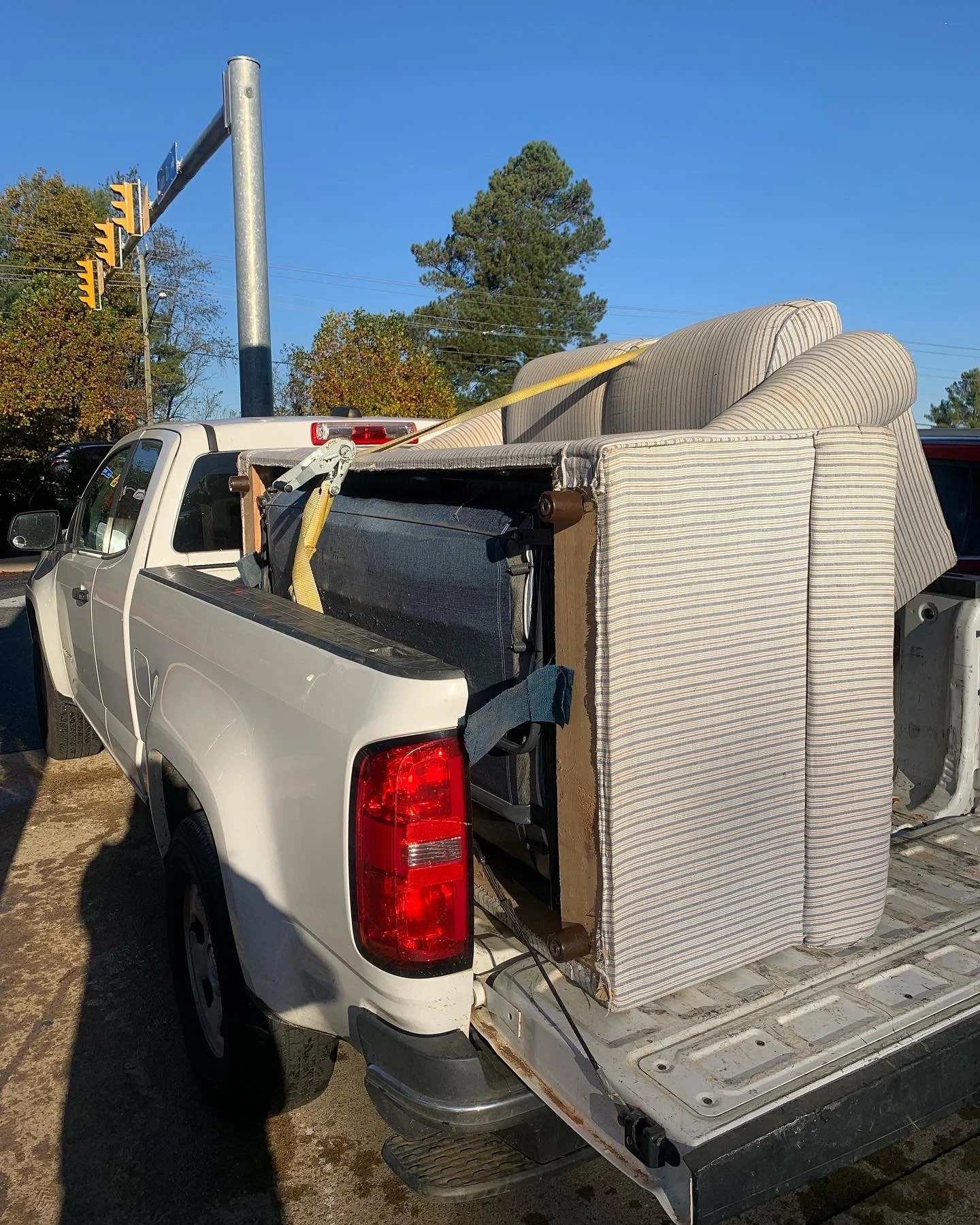 A white pickup truck with furniture loaded in the truck bed, including a striped sofa and other cushions, secured with yellow straps.