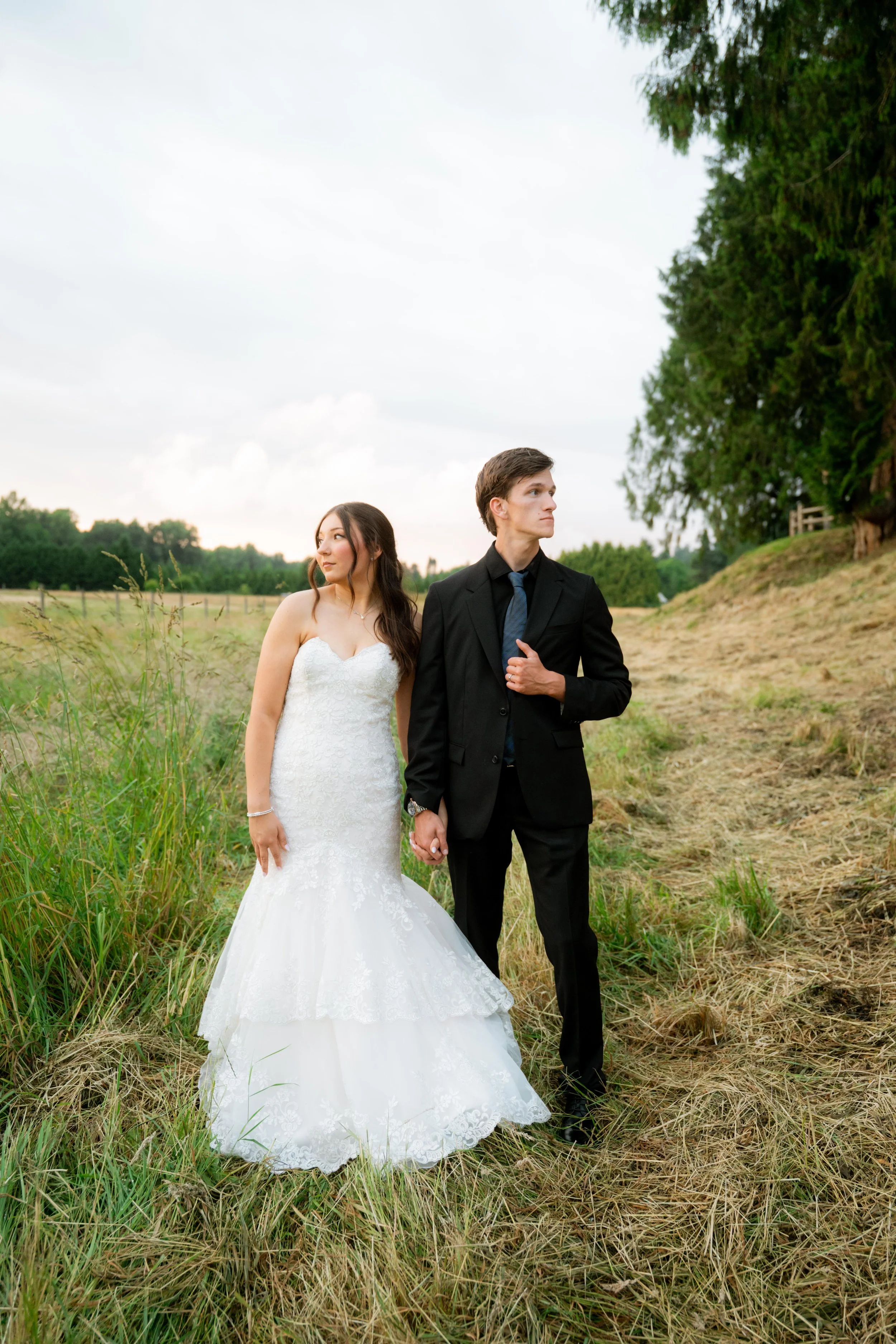 A bride in a white wedding gown holding hands with a groom in a black suit walk through a grassy field with trees in the background under a cloudy sky.