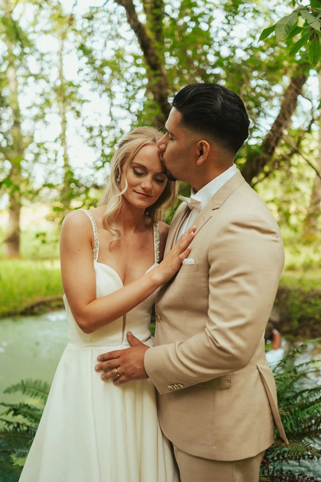 A bride and groom sharing an intimate moment outdoors, the groom kissing the bride on the forehead; lush green trees and a pond in the background.