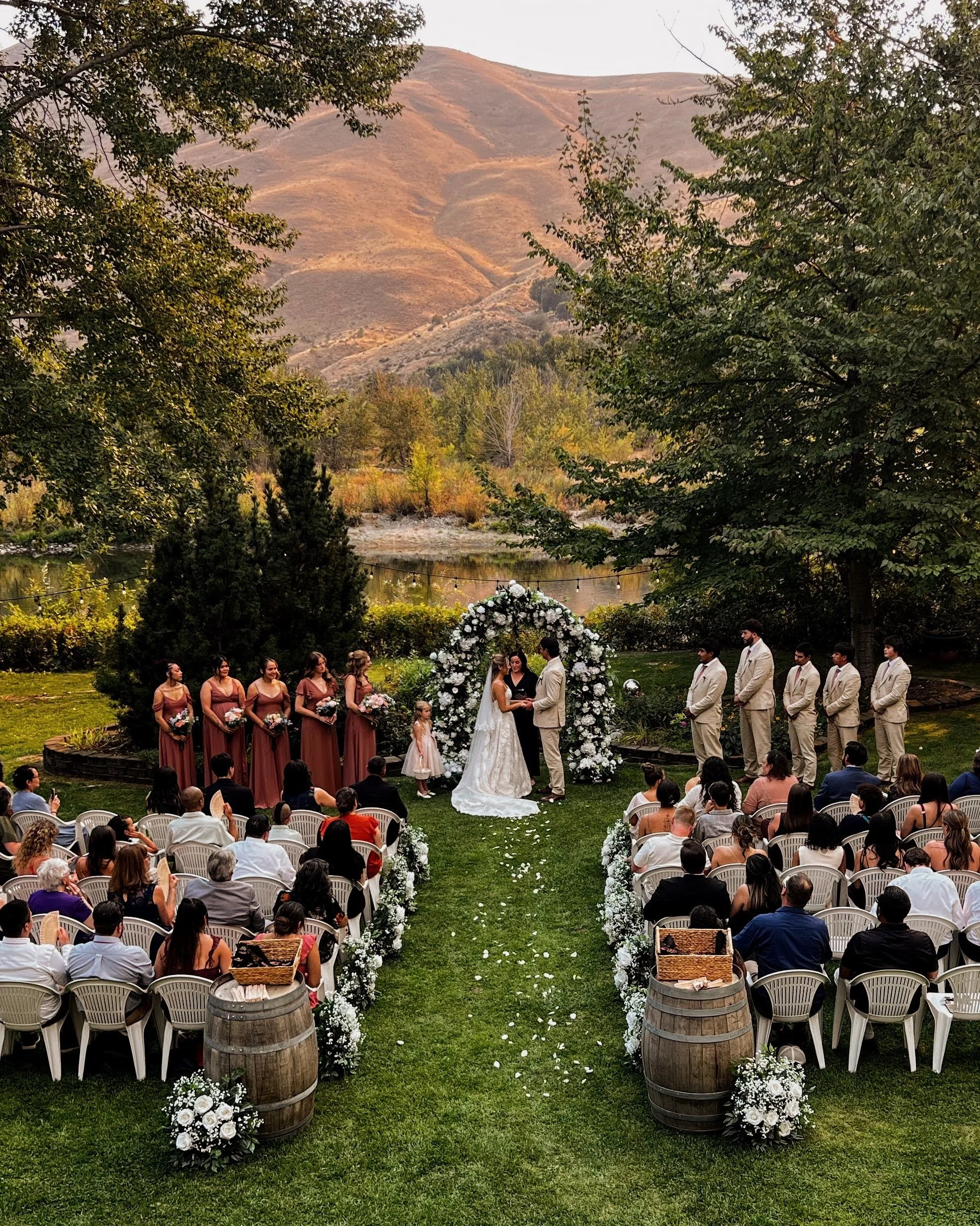 A wedding ceremony taking place outdoors with the couple exchanging vows under a floral arch, surrounded by bridesmaids in mauve dresses, groomsmen in beige suits, guests seated on white chairs, and scenic mountains and trees in the background.