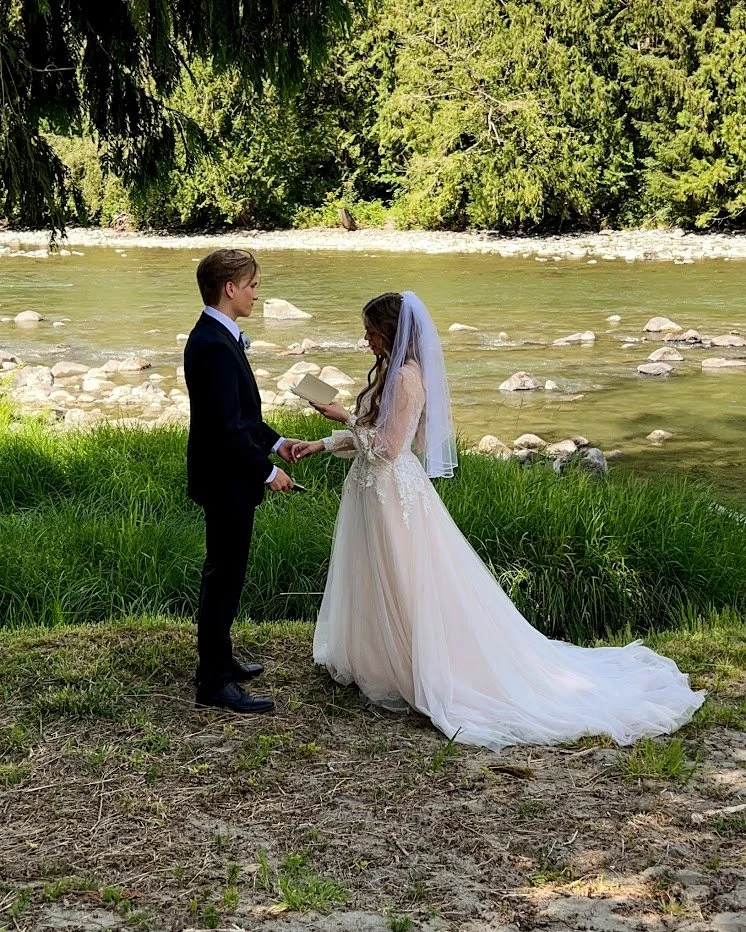 A bride and groom exchanging vows near a river, with lush green trees in the background.