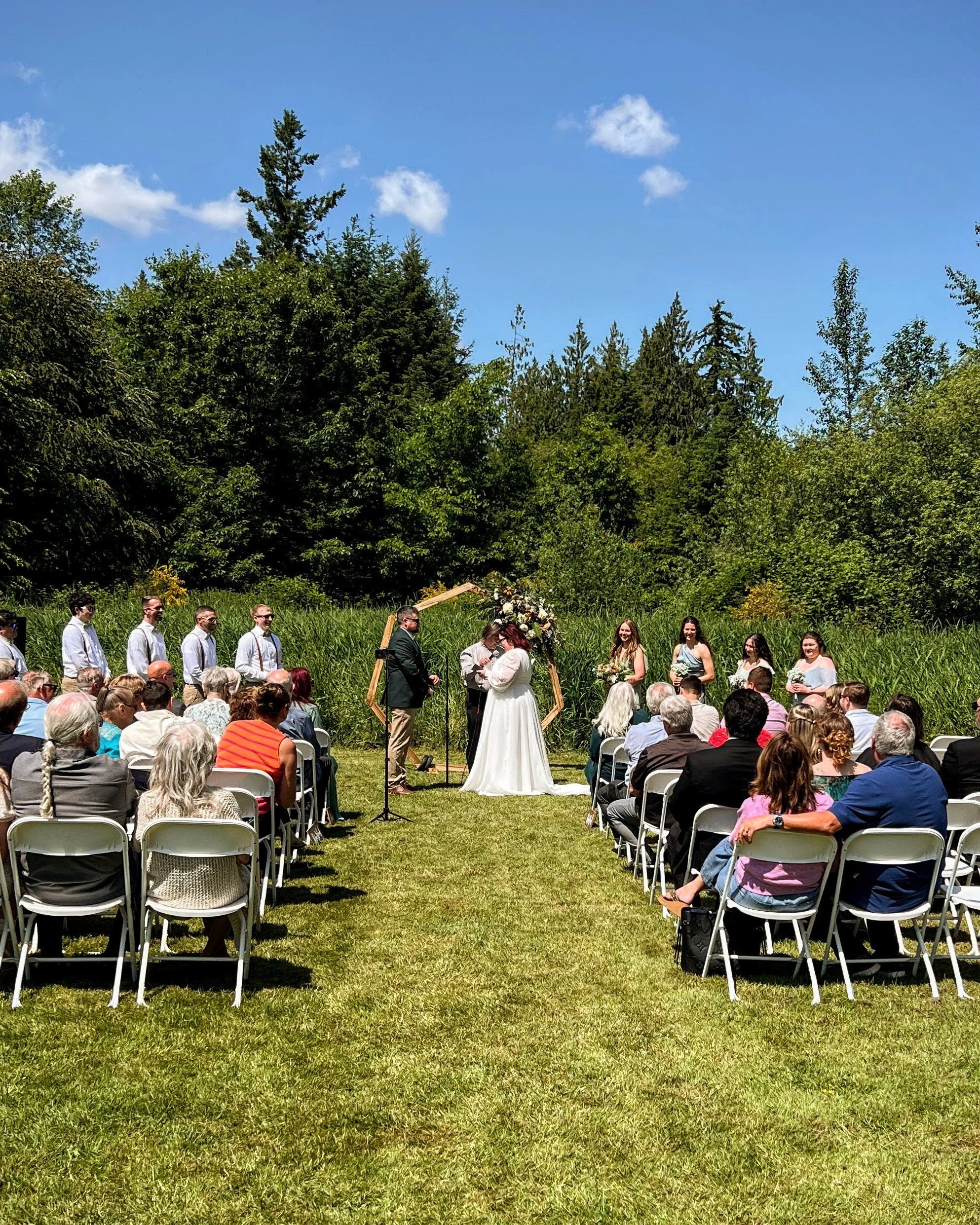 Outdoor wedding ceremony with bride and groom, officiant, and bridesmaids and groomsmen in natural greenery setting under a blue sky.