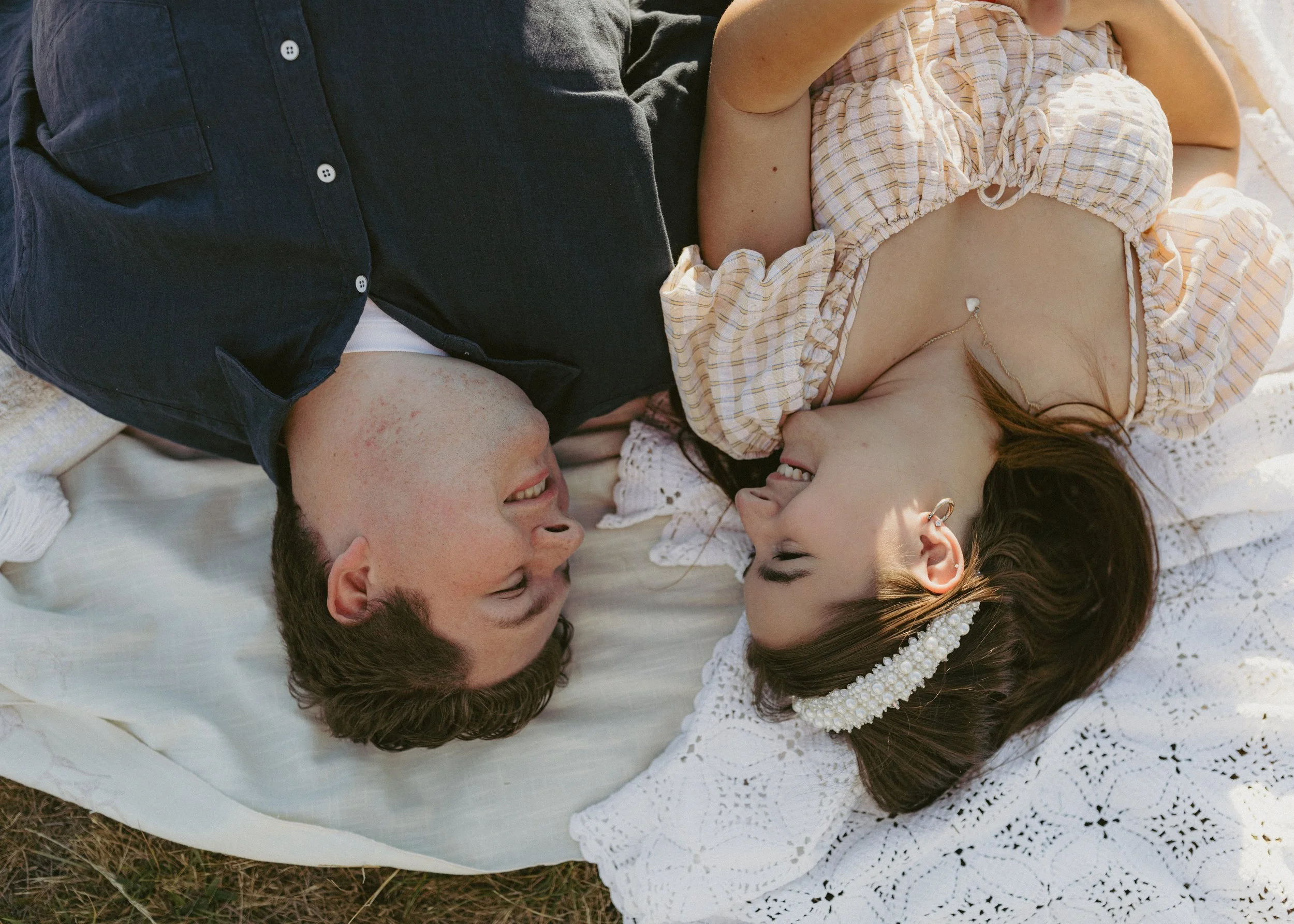A young couple lying on a white lace blanket, facing each other and smiling, outdoors in natural light.