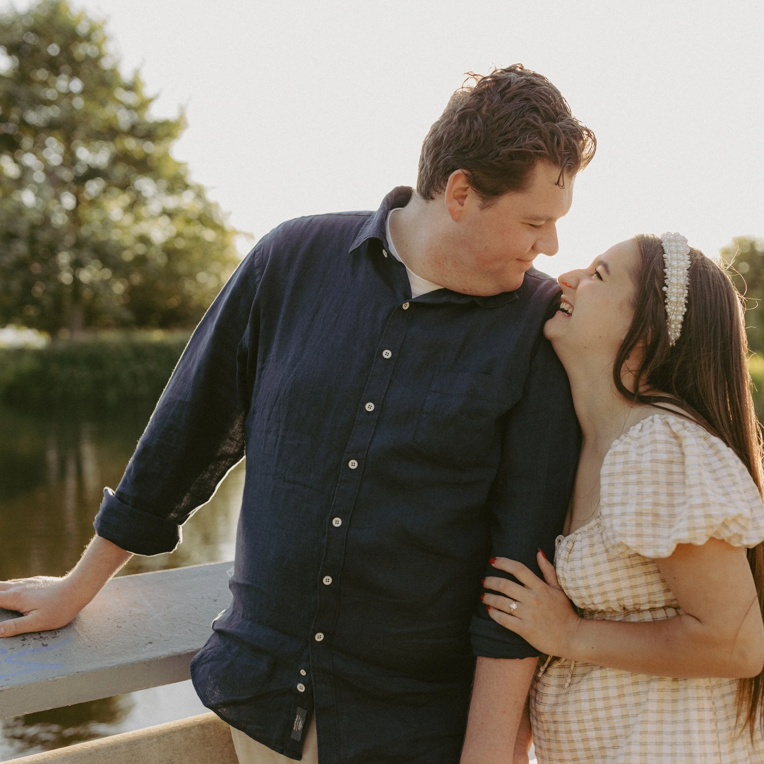 A couple standing close together on a bridge near a body of water, smiling and looking at each other at sunset, outdoors with trees in the background.