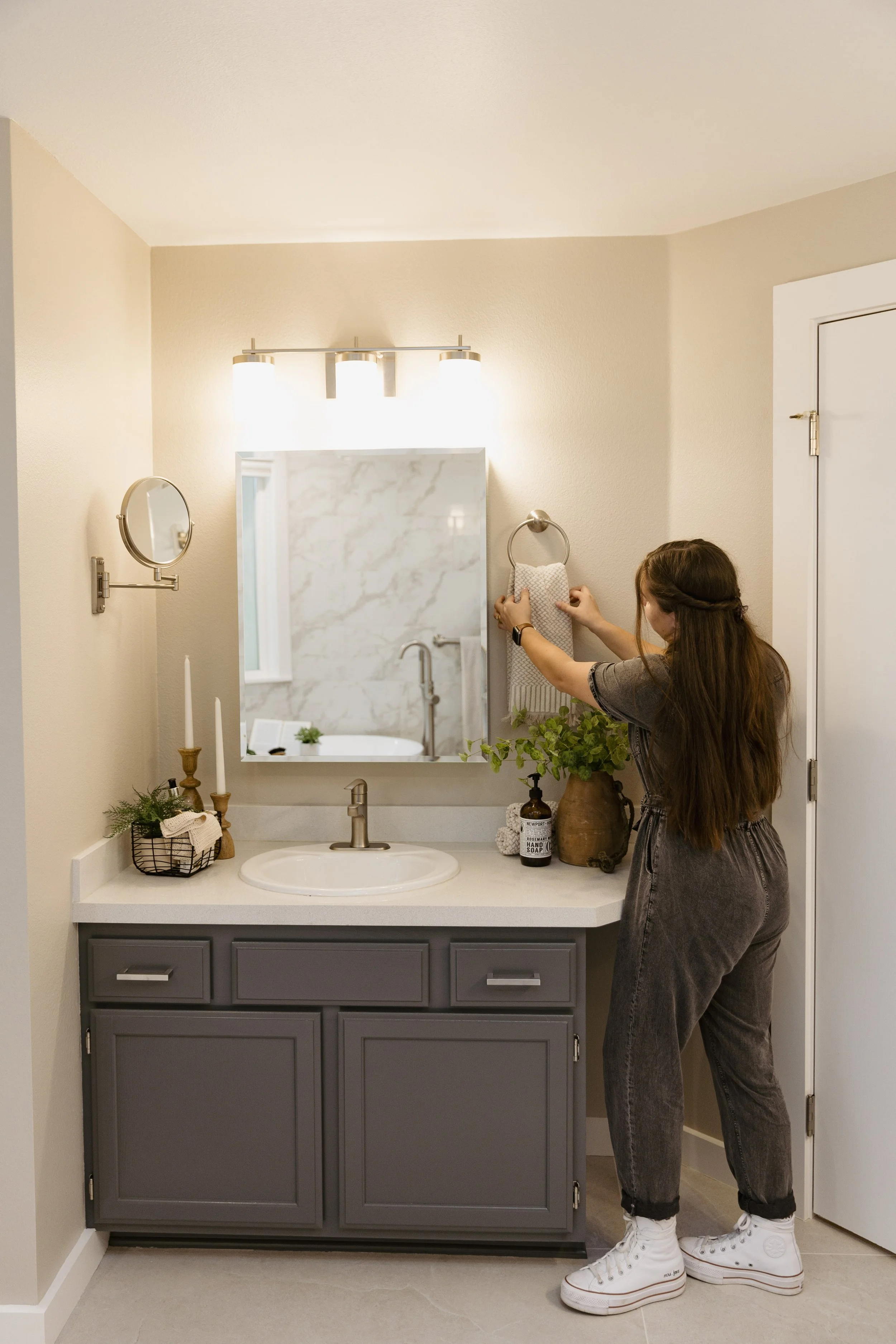 A woman decorating a bathroom vanity with a towel ring, mirror, and vanity with a sink. The bathroom has a mirror, wall-mounted light, and decorative items on the counter.