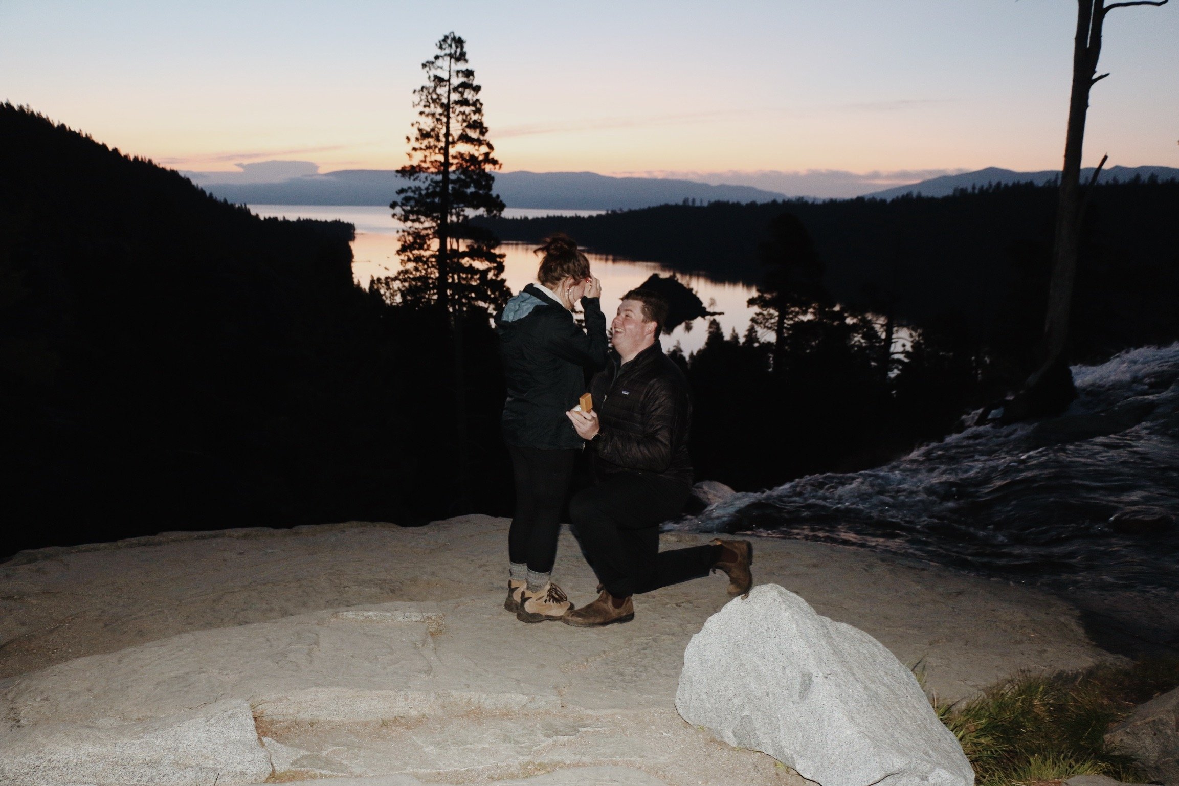 A man proposing marriage to a woman on a rocky ledge with a scenic lake and sunset in the background.