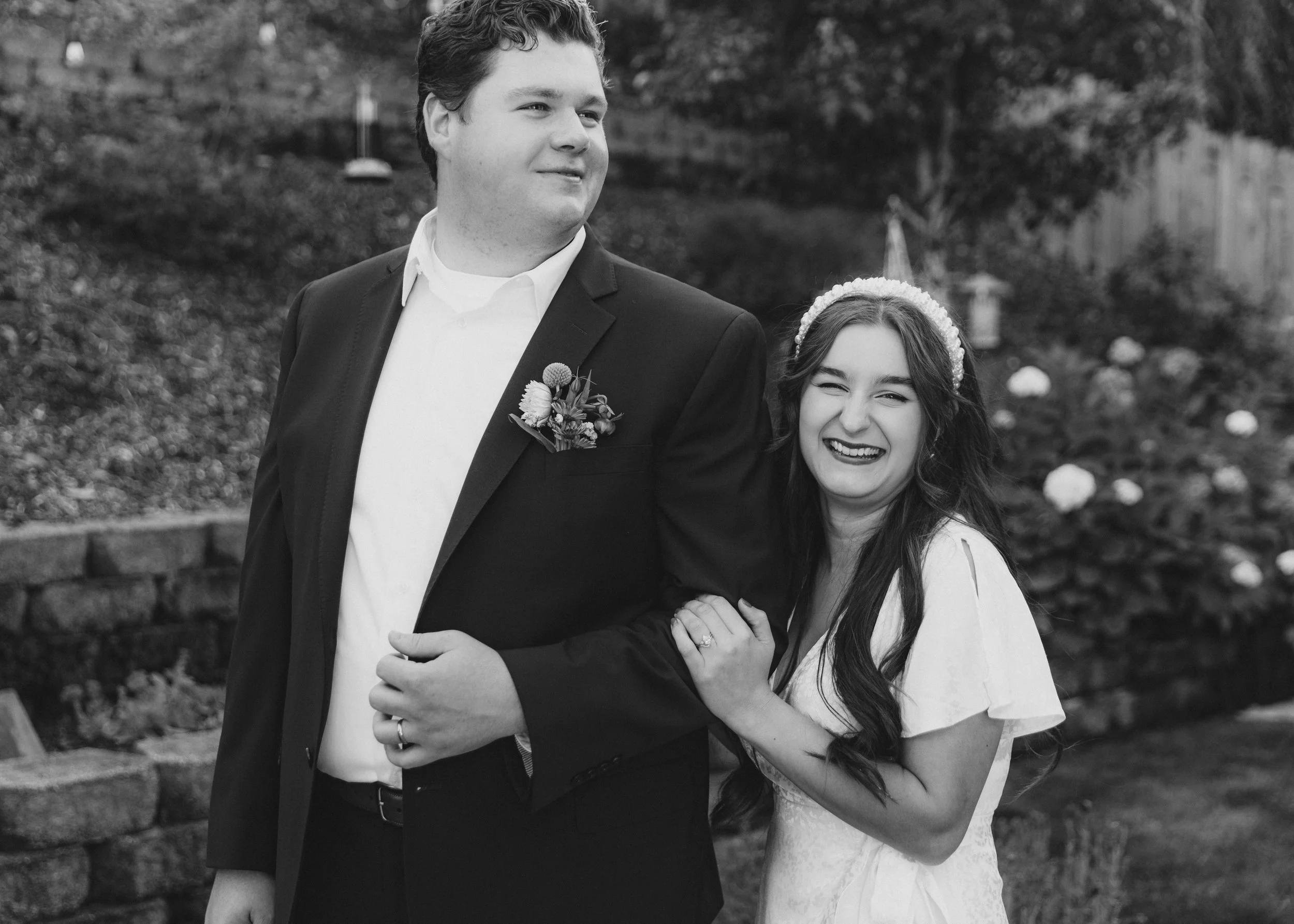 A happy couple at a wedding, the man in a suit with a boutonniere and the woman in a dress with a pearl headband, smiling and holding hands outdoors.