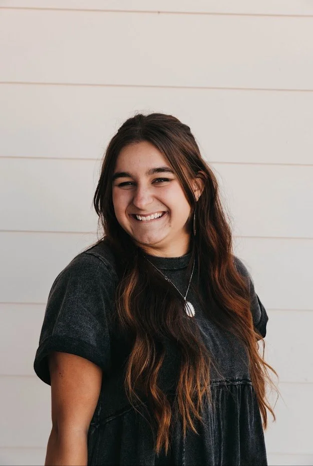 A young woman with long, wavy brown hair smiling and looking to her left, wearing a dark t-shirt and necklace, standing against a white shiplap wall.