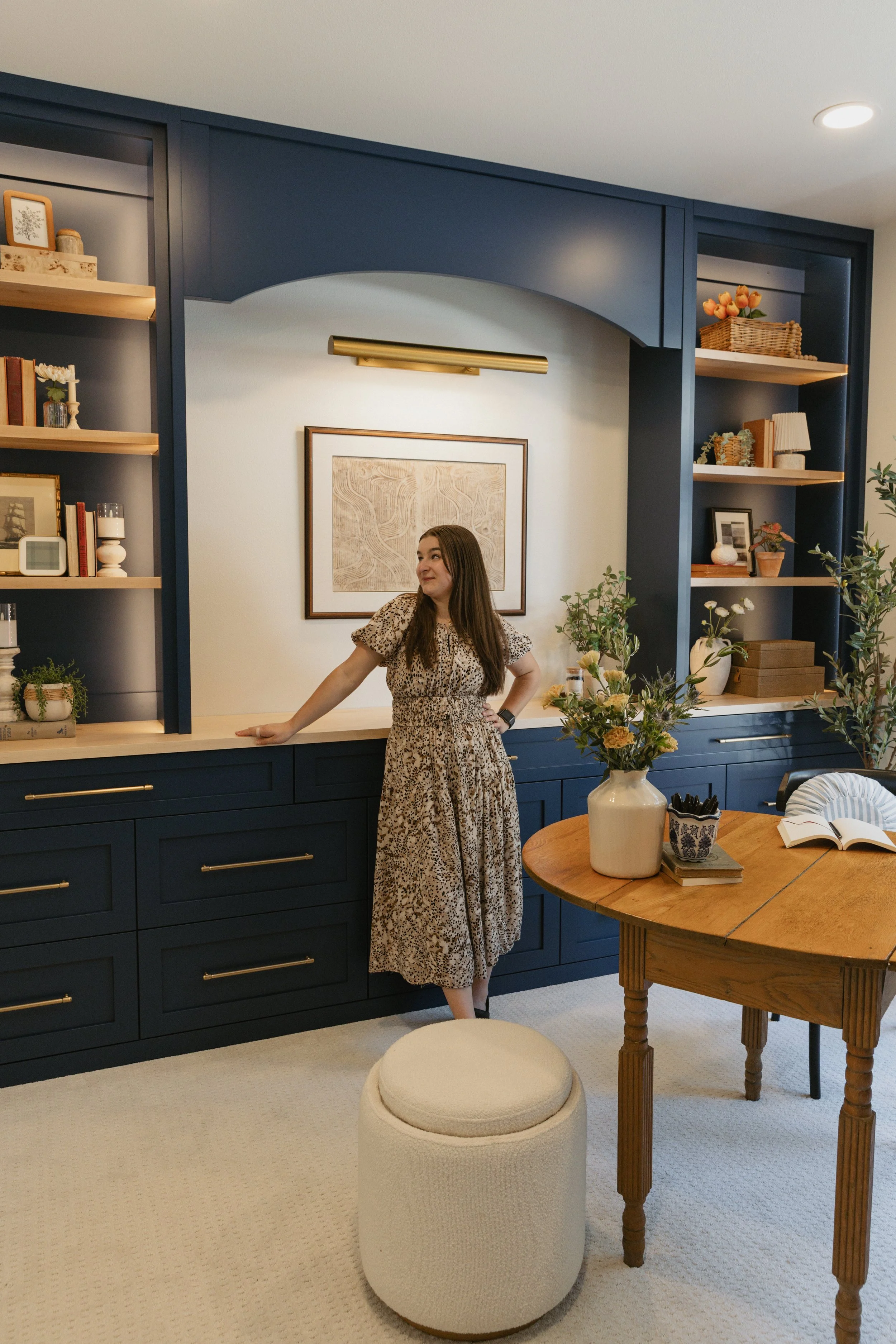 Woman in leopard print dress standing in a modern dining room with dark blue cabinets, a wooden dining table, a large vase of flowers, and built-in shelves decorated with books and decorative items.
