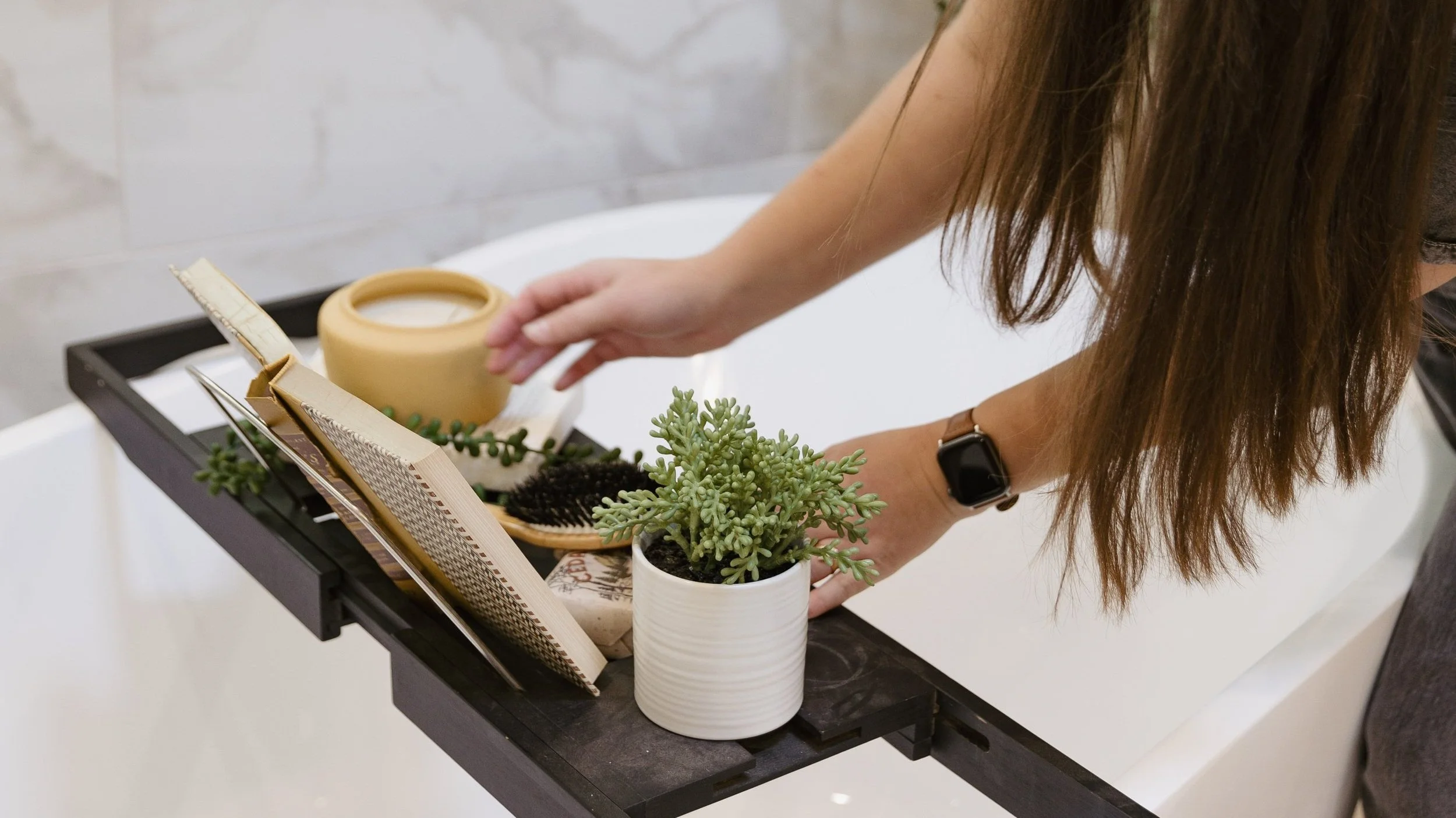Person adjusting a potted succulent plant in a bathtub with various household items, including a magazine, a hairbrush, and a decorative ceramic bowl.