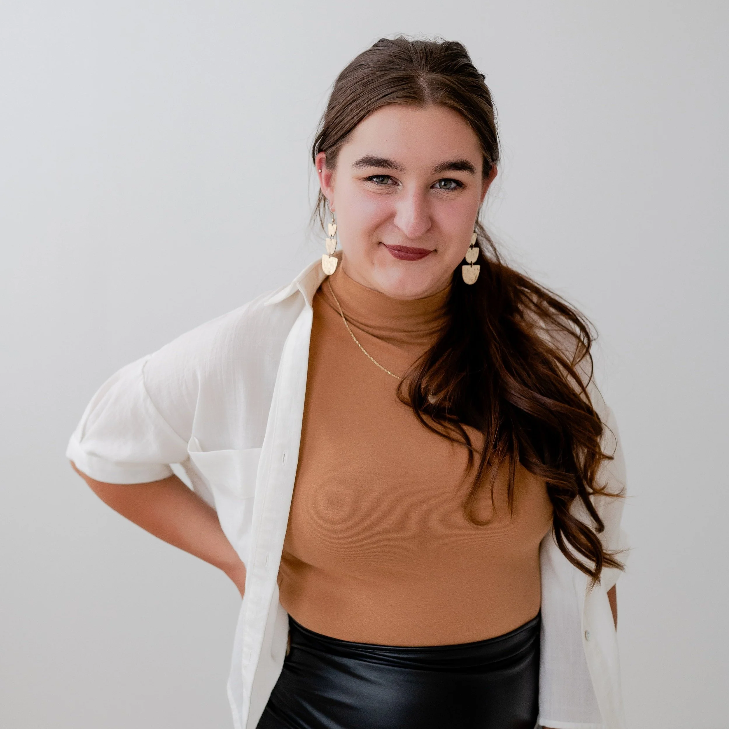 A young woman with long brown hair, wearing a tan turtleneck, white shirt, black shiny skirt, and gold dangle earrings, smiling at the camera against a plain white background.