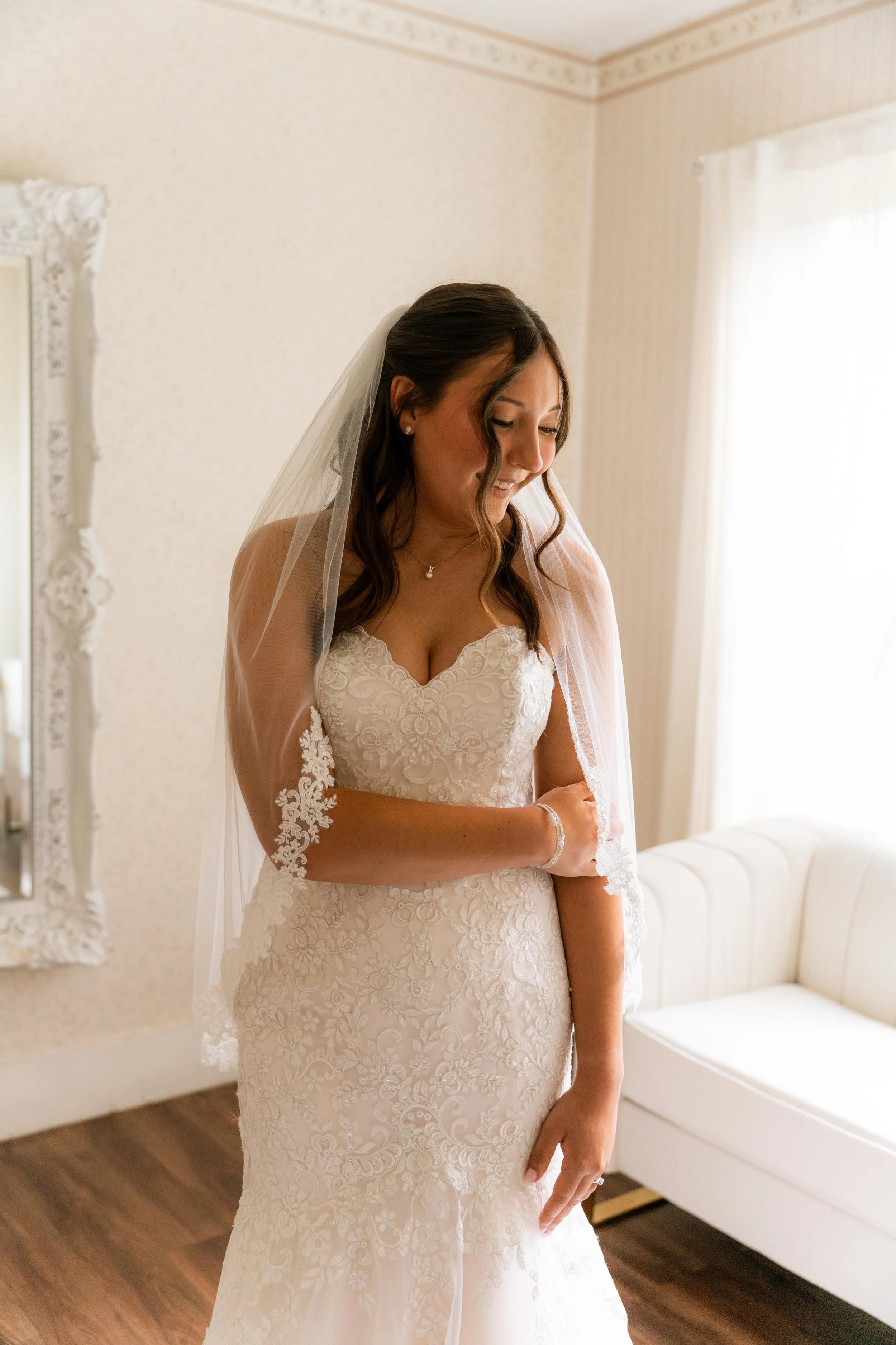 A bride in a detailed lace wedding gown with floral embroidery, a veil, and pearl earrings, standing indoors by a window with sheer curtains, smiling gently.