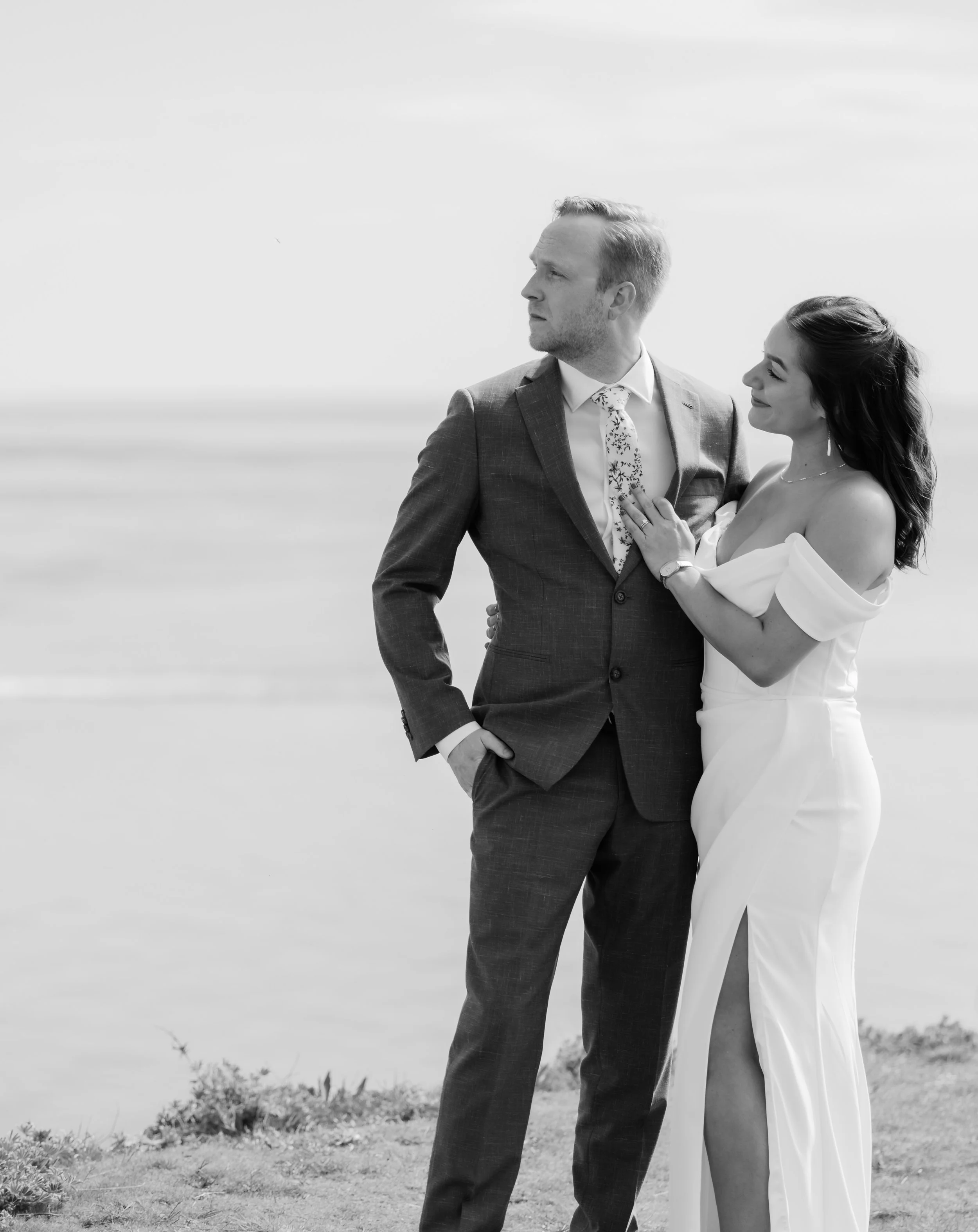 A man in a suit and a woman in a wedding dress standing outdoors on a beach, looking into the distance.