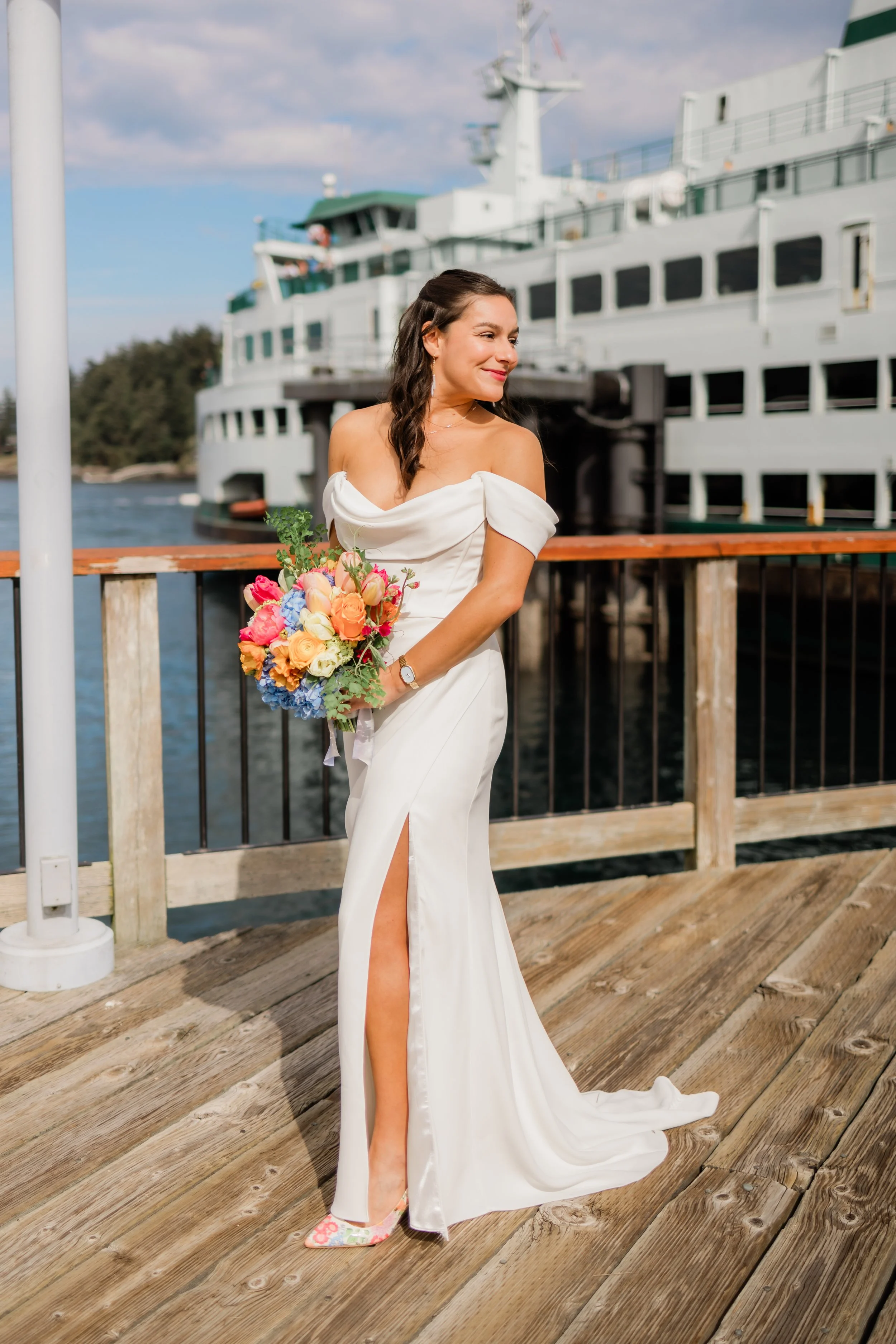 A woman in a white wedding dress holding a colorful bouquet of flowers standing on a wooden dock with a boat in the background.