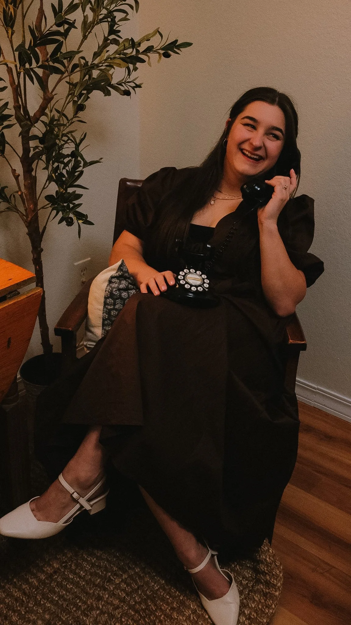 A woman sitting on a wooden chair, smiling and talking on a black rotary phone. She is wearing a brown dress and white high heels, with a large indoor plant nearby.