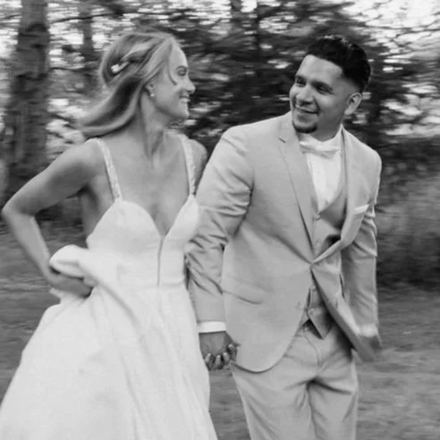 Black and white photo of a happy couple in wedding attire walking outdoors, holding hands, smiling, surrounded by trees.