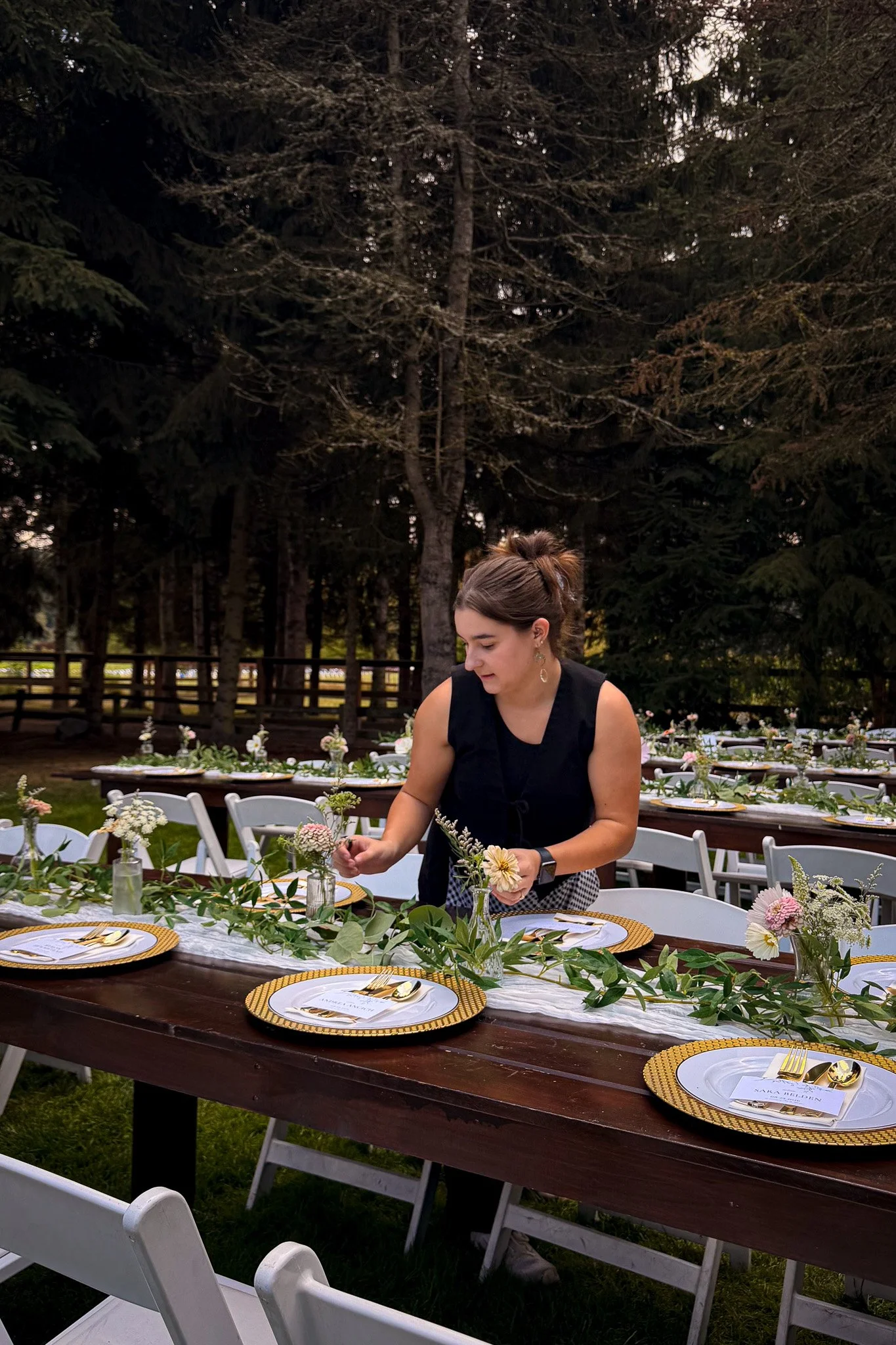 Savannah Fox, lead planner of Made Merrily, arranging floral centerpieces on a dinner table outdoors, with a wooded background and white chairs.