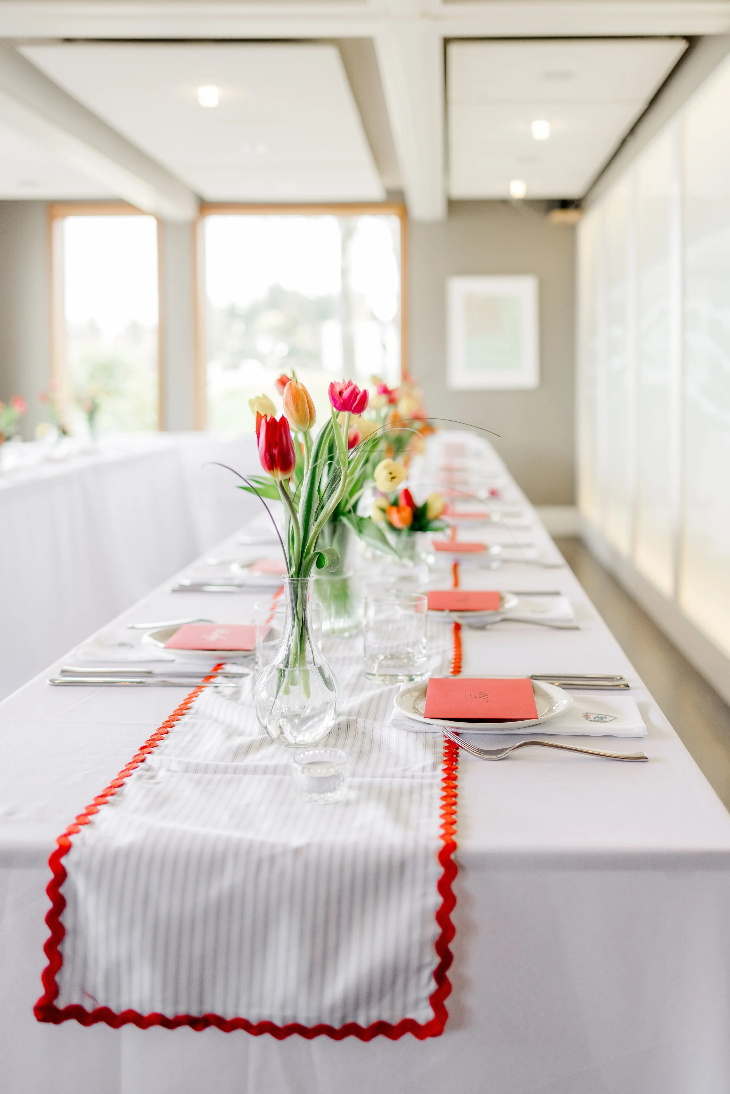 A long rectangular dining table decorated with a striped white table runner with trim. The table is set with white plates, silverware, pink and orange folded napkins, and small votive candle holders. A glass vase with multicolored tulips is placed in the center of the table. The room has large windows allowing natural light and some framed artwork on the walls.