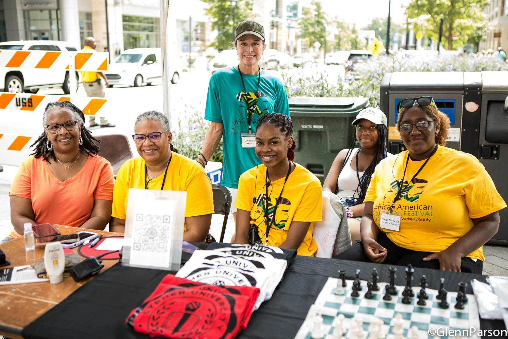 Group of women sitting at a table outdoors at the Africa American Cultural Festival in Wake County. The table has T-shirts, chessboard, and other items. One woman stands behind the seated women.