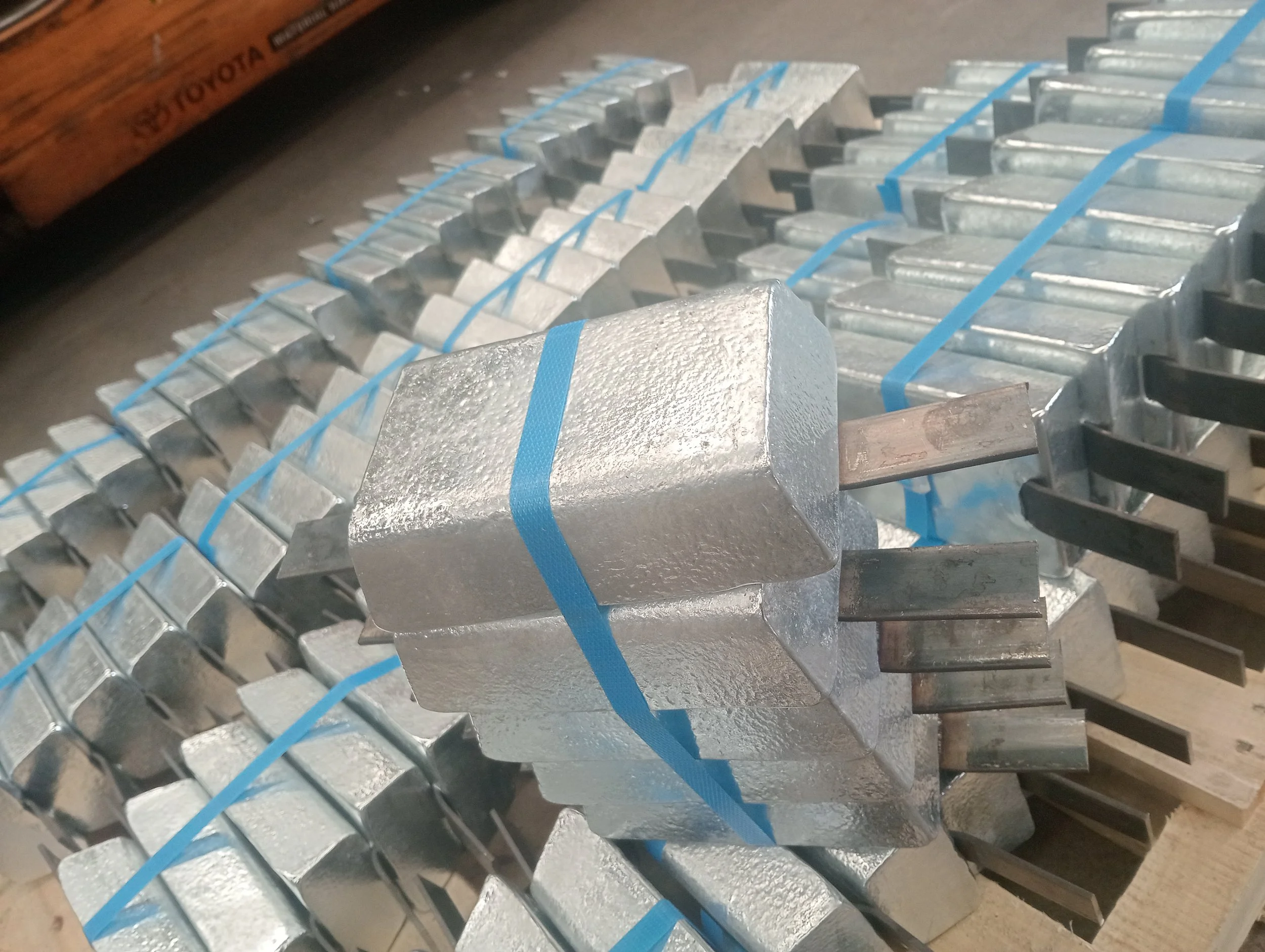 Stack of metal ingots bound with blue straps, with one ingot in the foreground showing a protruding metal rod.