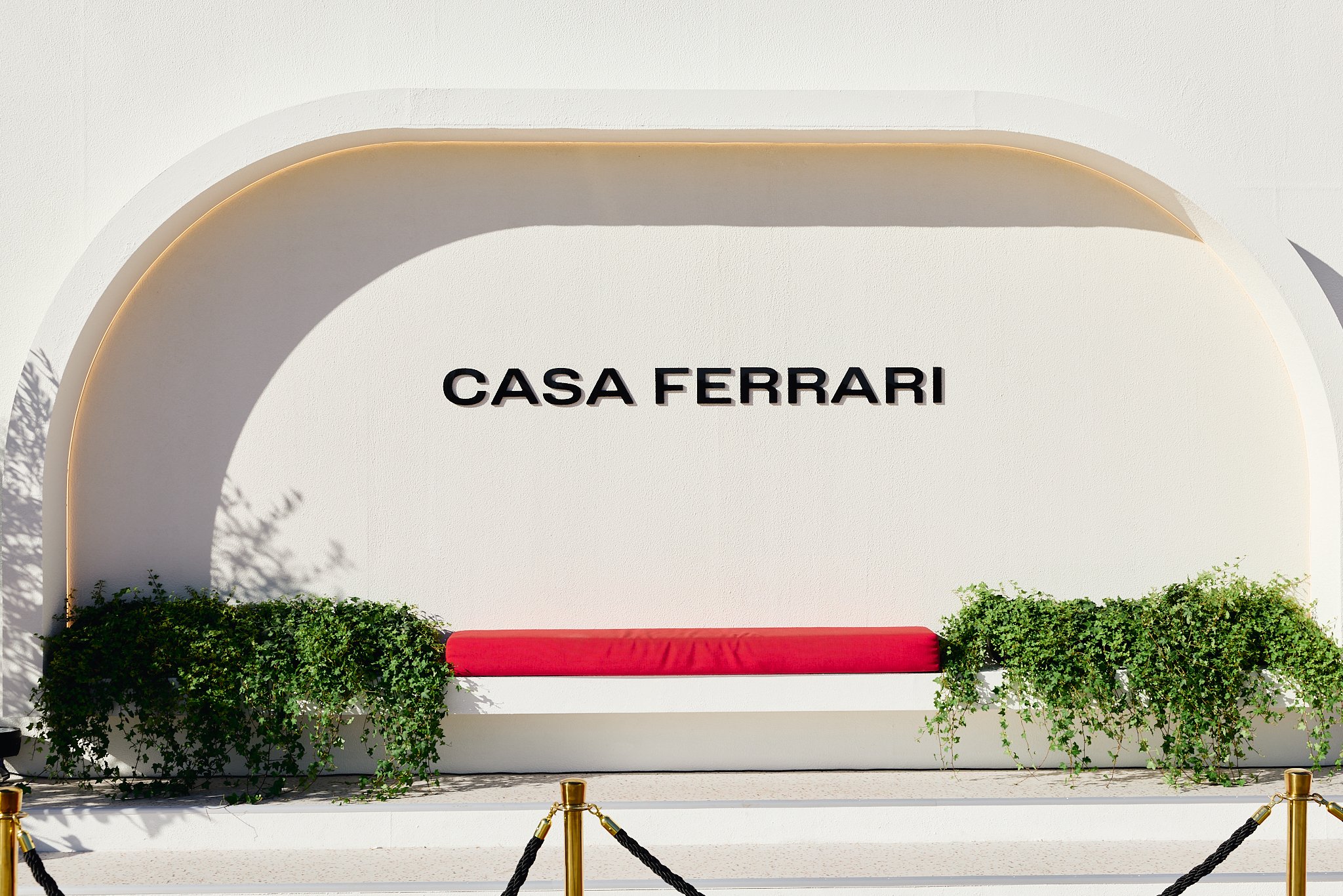 Front view of Casa Ferrari entrance with a white wall, black text, green bushes, and a red bench, roped off with black and gold barriers.