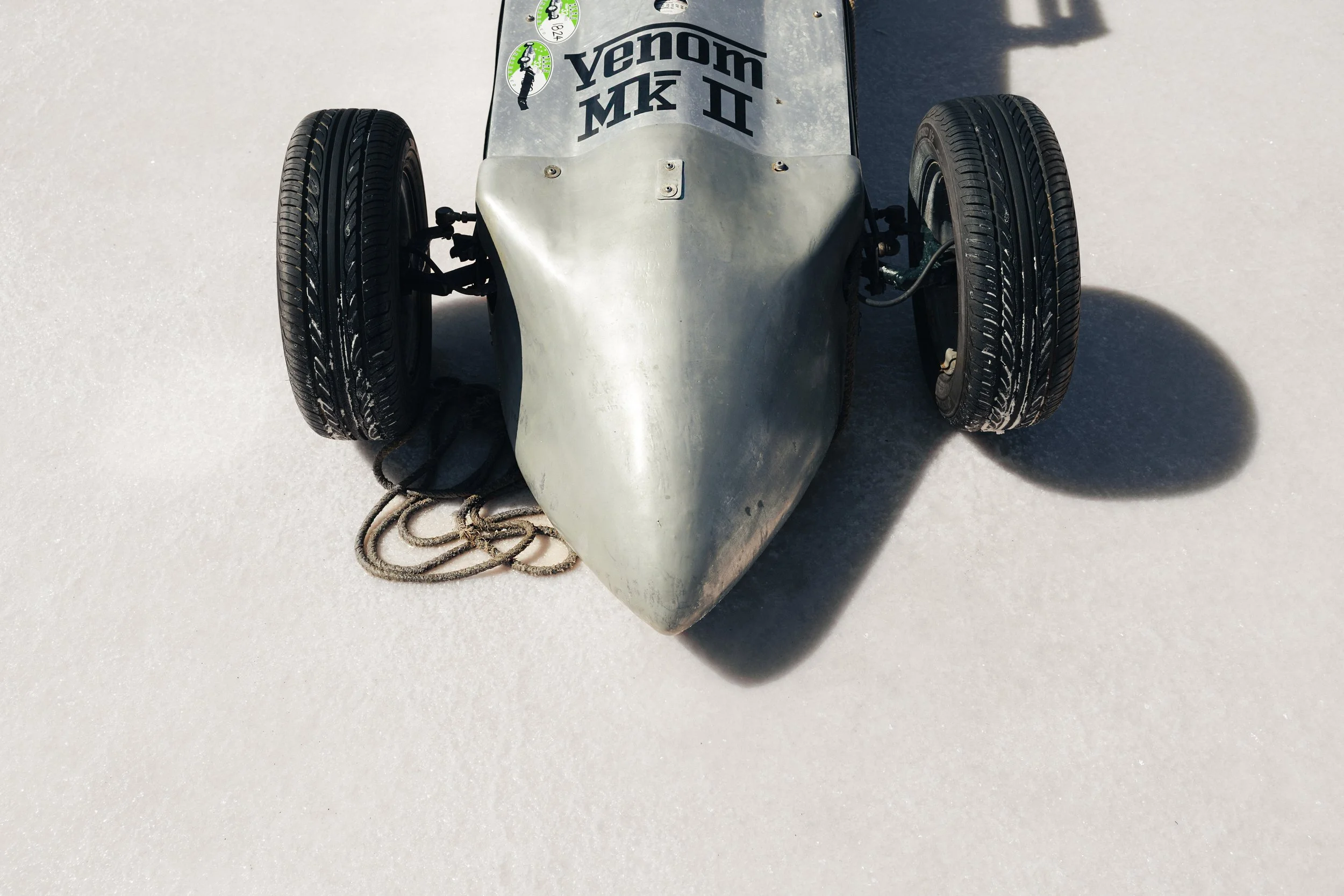 A silver vintage race car with the Kenora MK II logo on the front, black tires, and a rope lying on the white surface underneath it.