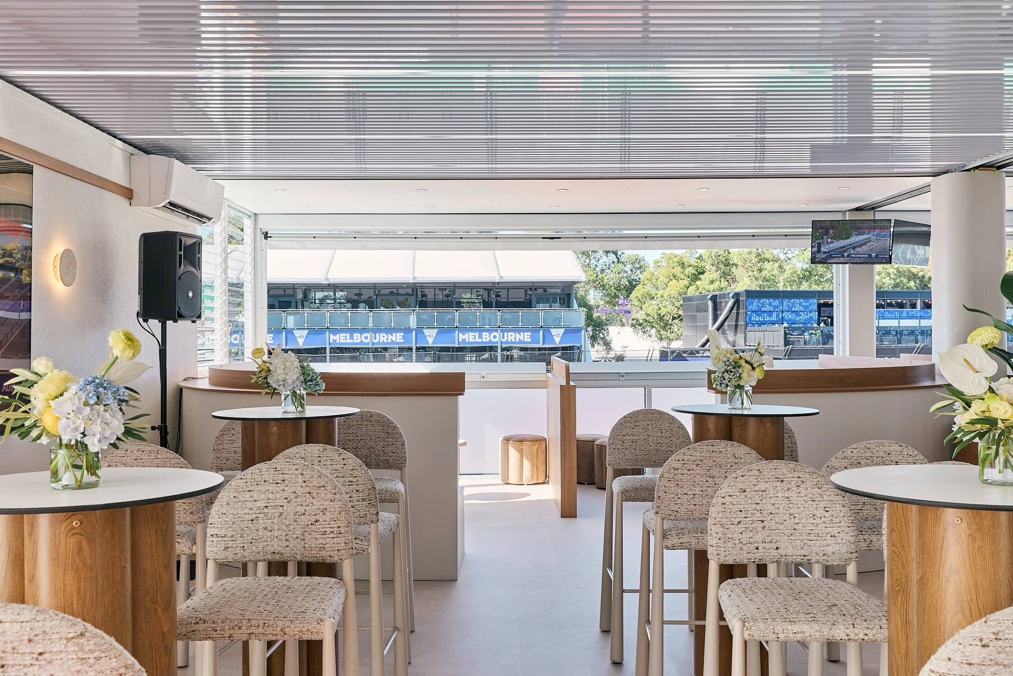 Interior of a modern lounge area with round tables decorated with vases of flowers, beige upholstered chairs, and large open windows showing a view of the Melbourne racecourse outside.