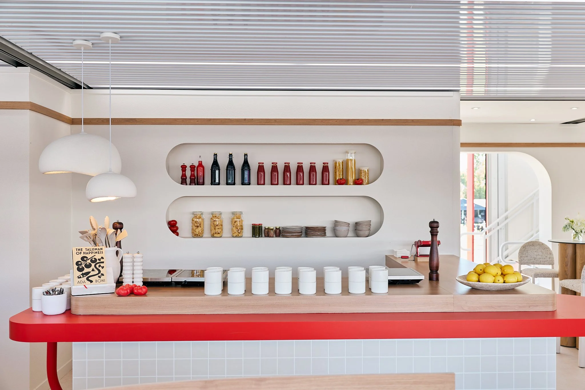 Modern cafe or kitchen counter with white and red decor, jars of pasta, tomatoes, and lemons, shelves with bottles and jars, and white pendant lights hanging from the ceiling.