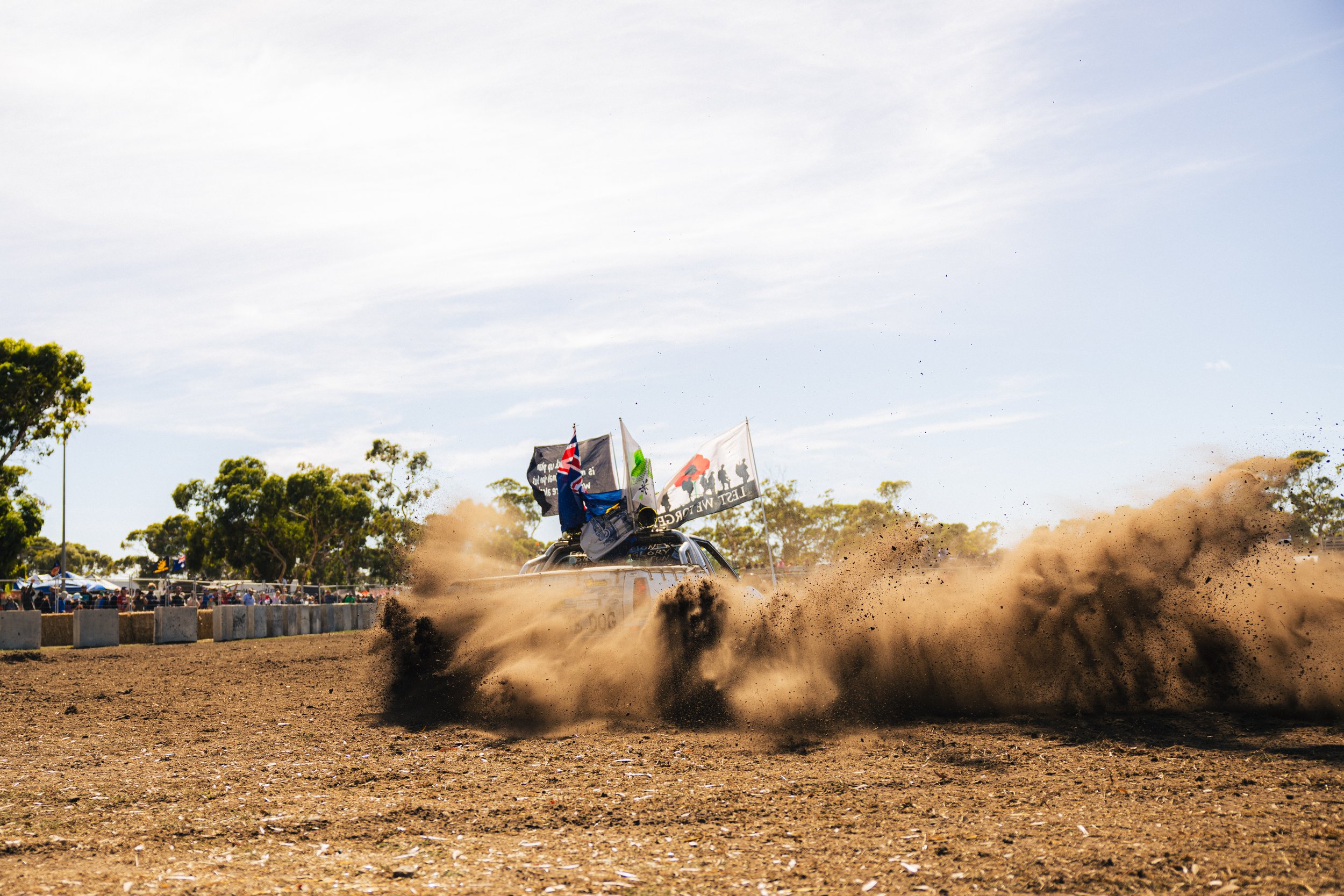 A vehicle kicking up dust on a dirt track during a race, with flags mounted on it and spectators in the background.
