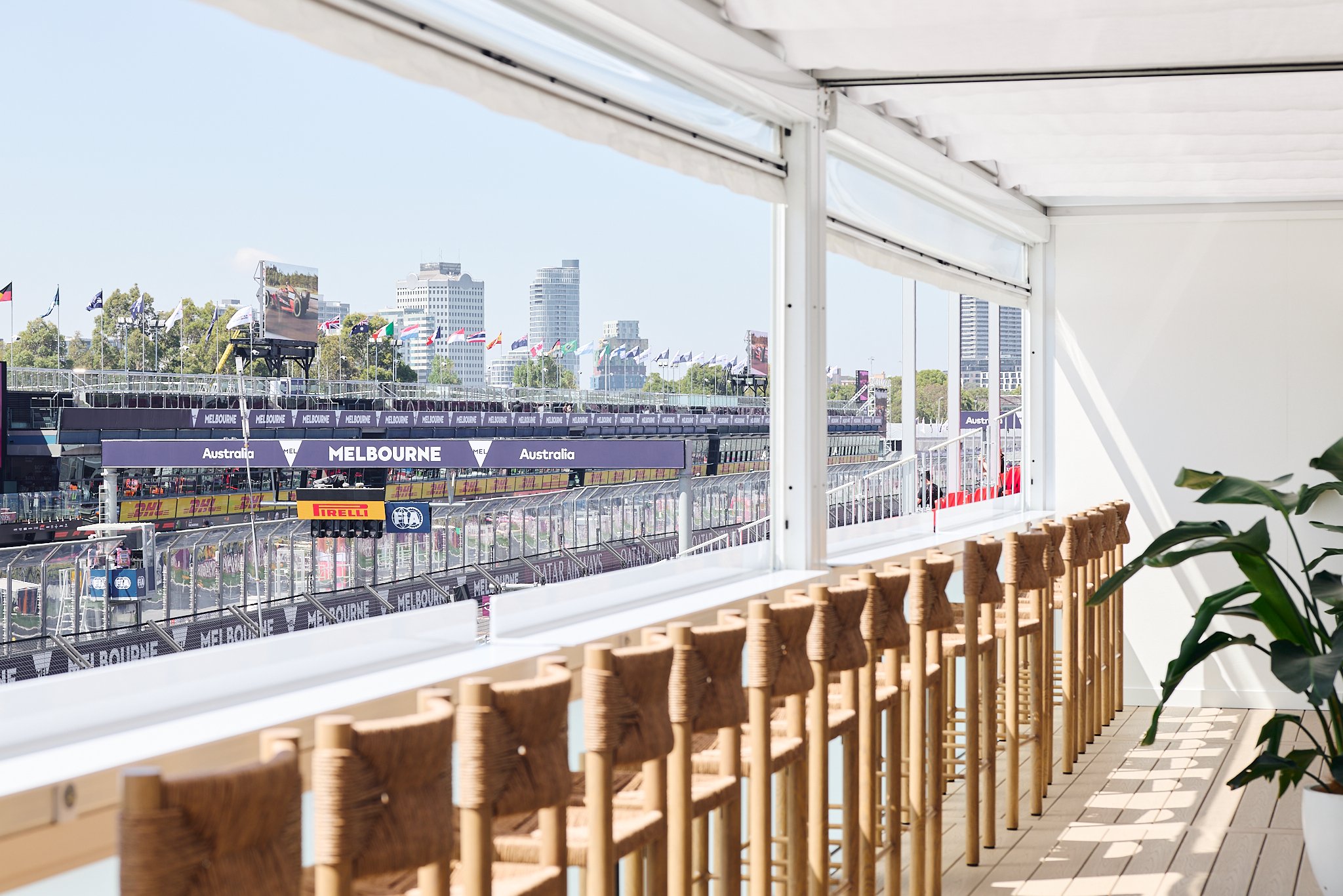 View of a Formula 1 race track from a balcony with wooden chairs, flags, and tall buildings in the background.