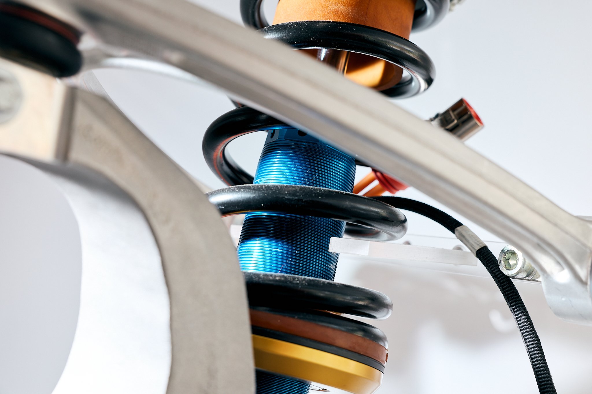 Close-up of a motorcycle rear suspension showing a blue coil shock absorber with a metal surrounding frame and some wiring.