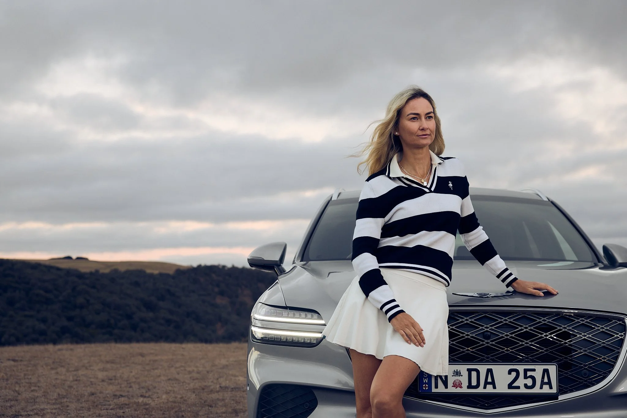 A woman with blonde hair standing beside a silver luxury car in an open field under a cloudy sky.