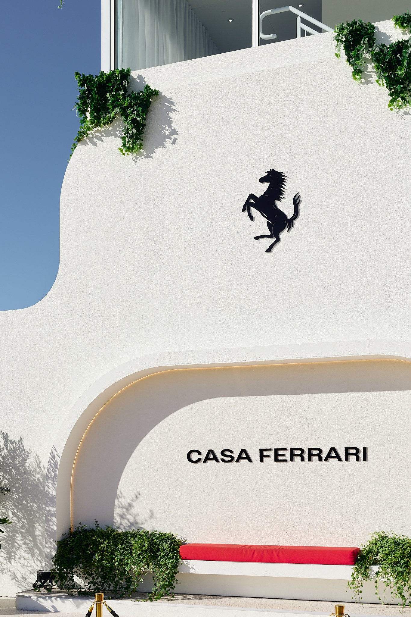 Modern white building with a black Ferrari logo and the words 'Casa Ferrari' on the front, with a curved architectural feature and greenery, under a blue sky.