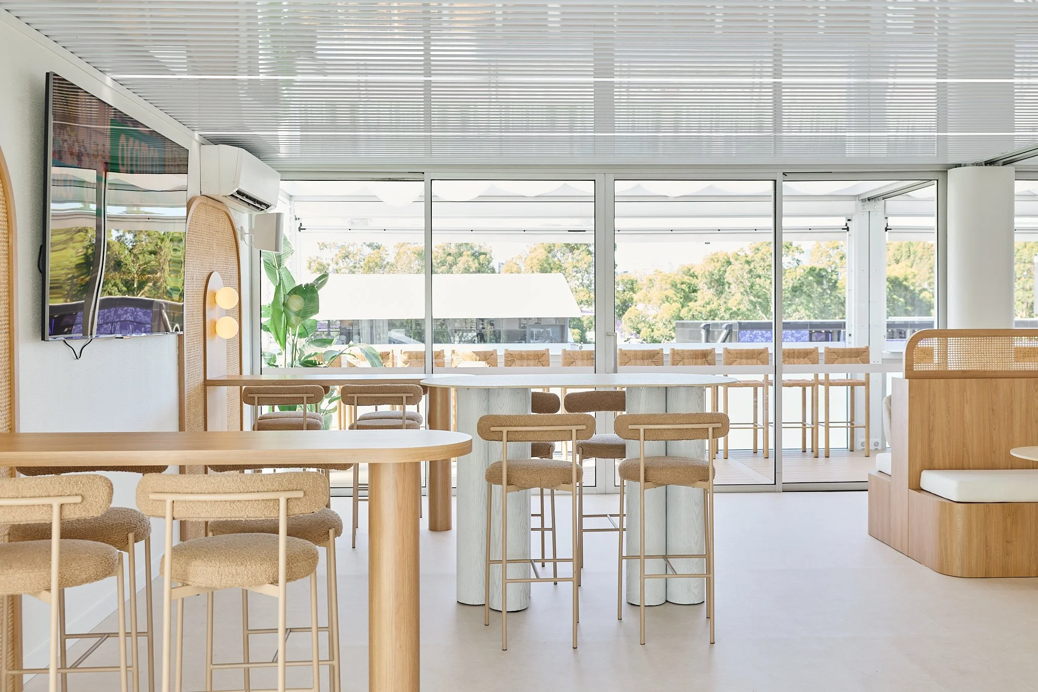 Modern, minimalist cafe or restaurant interior with light wood furnishings, bar stools, and large windows offering a view of greenery outside.