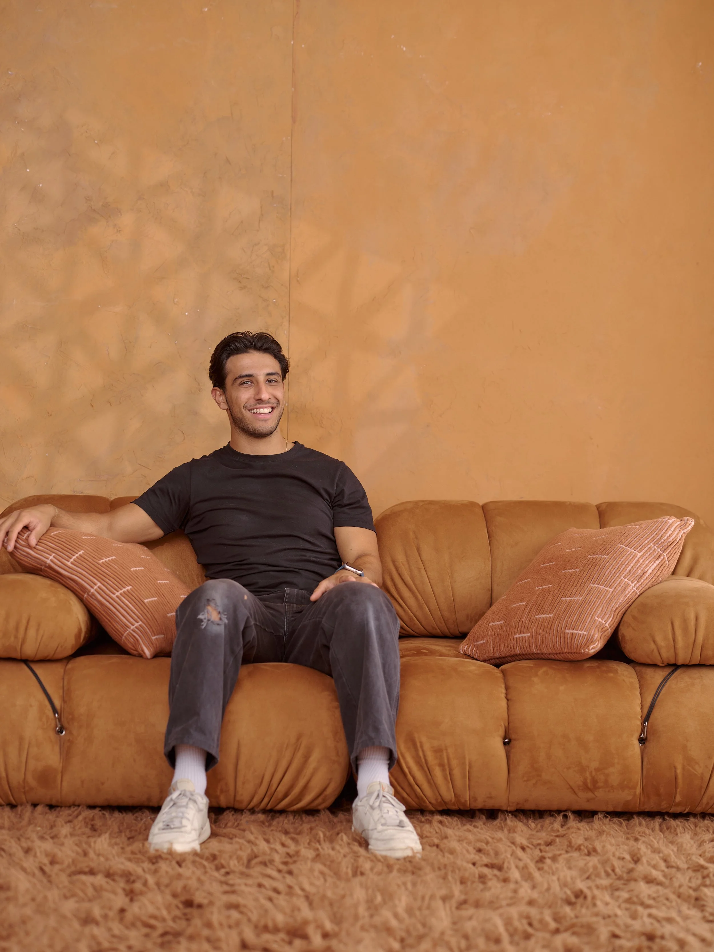 A young man with dark hair, wearing a black t-shirt, ripped jeans, and white sneakers, sitting on a shaggy brown carpet on a retro orange sofa with throw pillows, smiling at the camera with beige textured walls in the background.