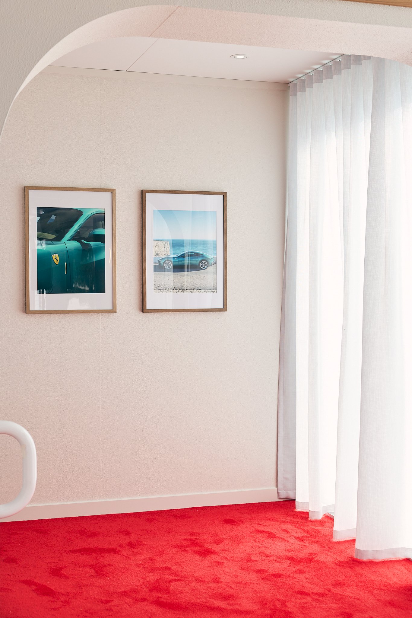 Interior of a room with a red carpet, white walls, and white curtains. Two framed photographs of cars are hanging on the wall, one showing a close-up of a car's exterior and the other a side view of a car near the ocean.