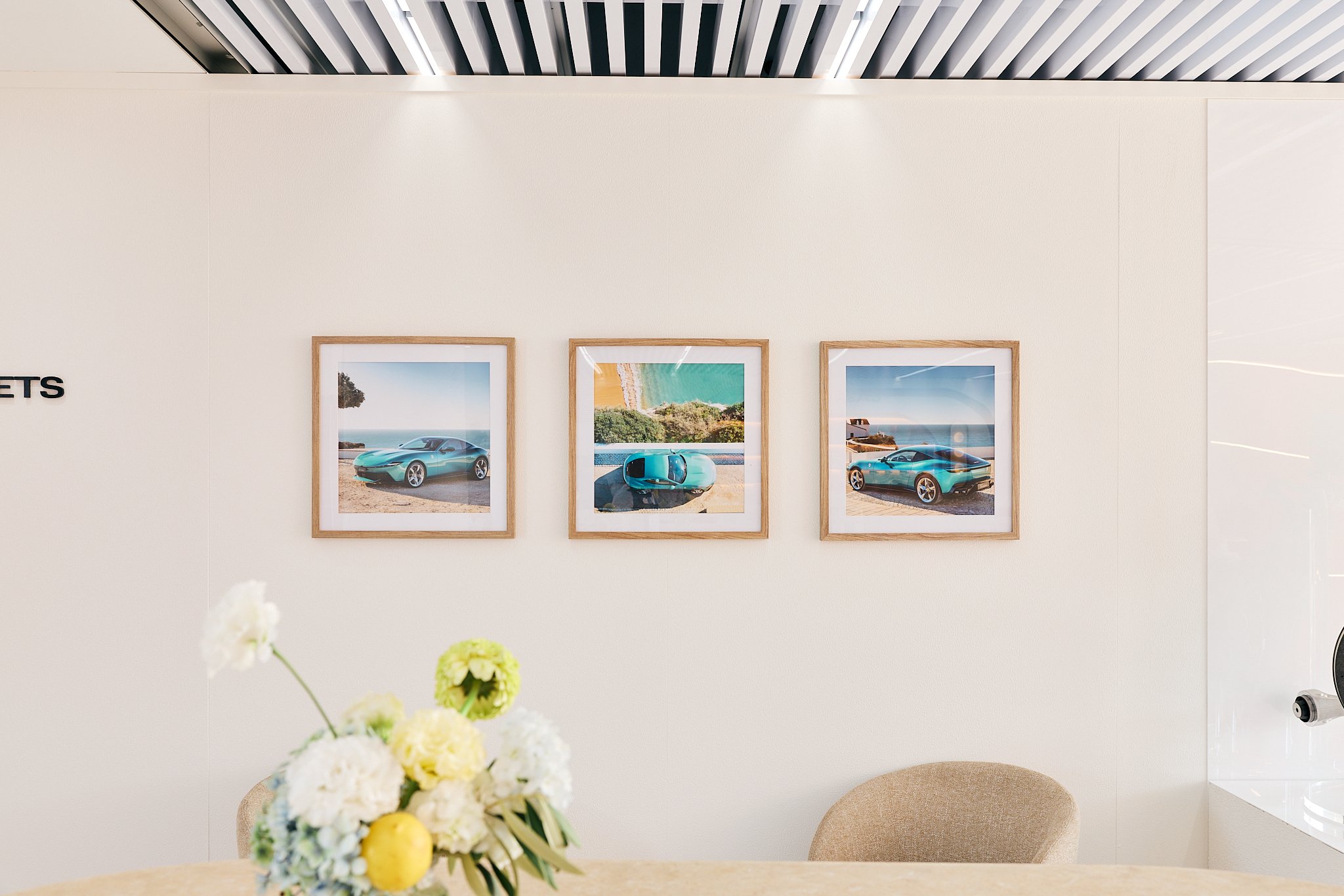 Three framed photos of teal cars in a bright, modern indoor space with a white wall and a beige chair and table with a flower arrangement.
