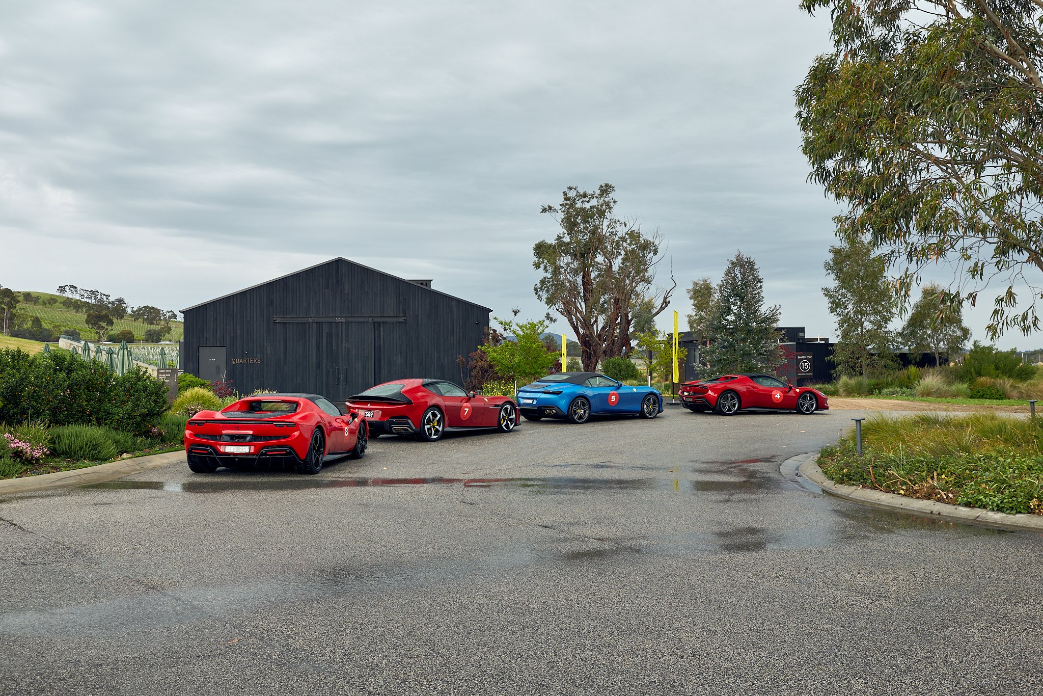 Five luxury sports cars parked on a wet driveway near a modern black shed surrounded by greenery and trees.