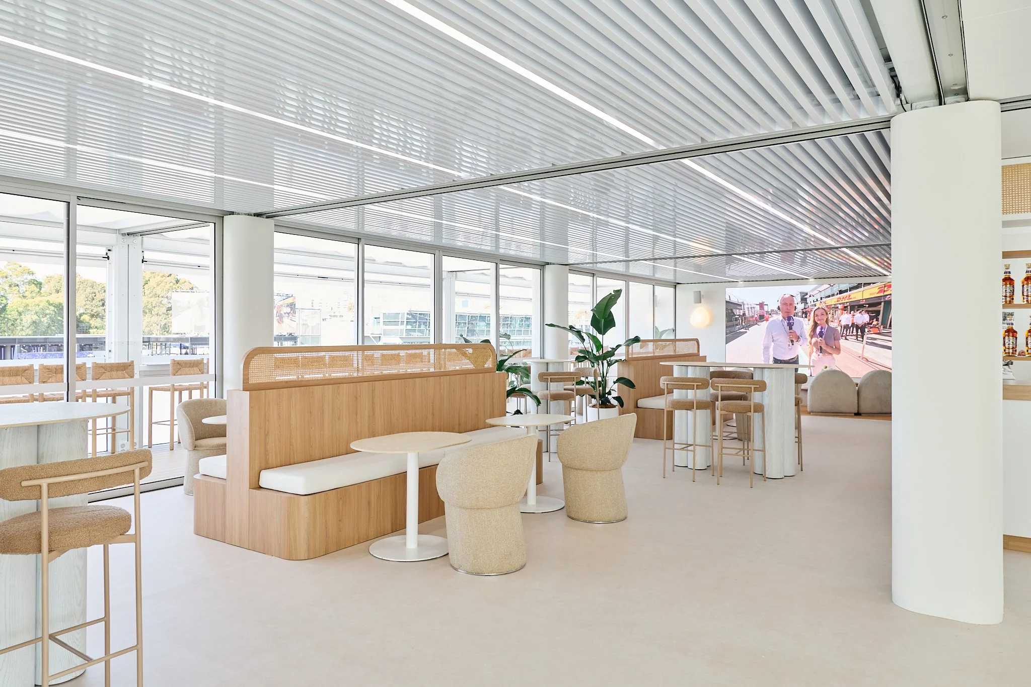 Modern indoor lounge area with beige chairs, white tables, wooden partition, potted plants, and a large screen on the wall showing two news reporters.