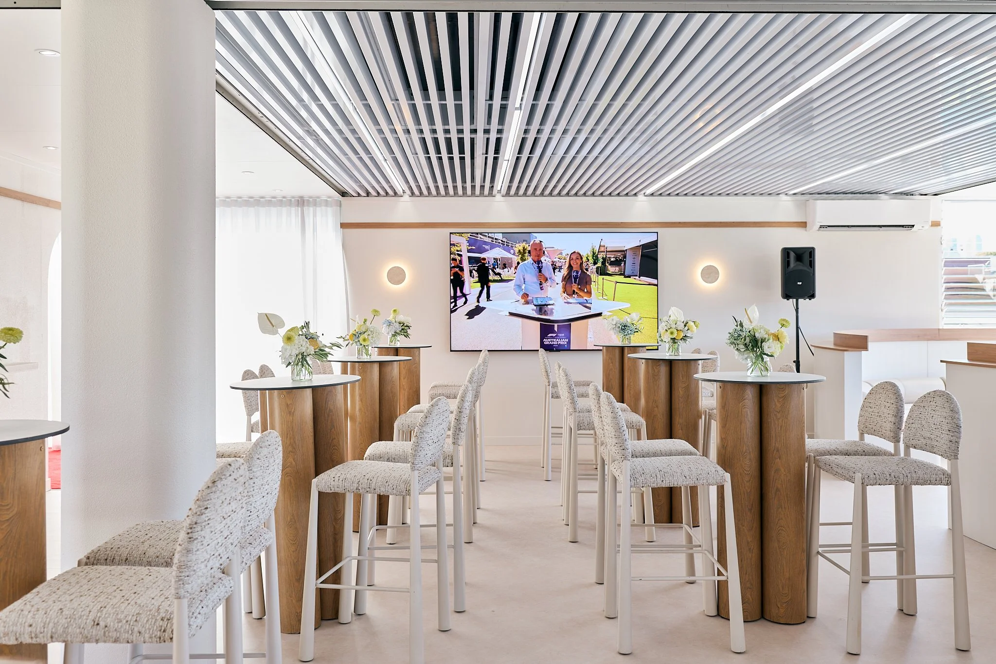 Indoor event space with high tables decorated with white flowers, chairs, a large screen on the wall, and a speaker, set up for a presentation or gathering.
