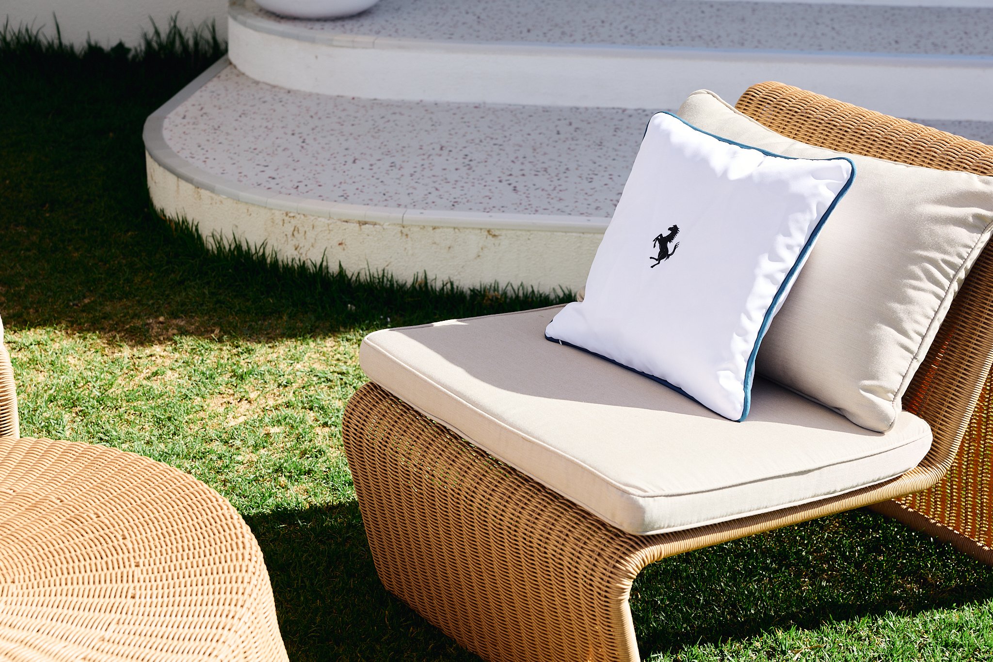 Outdoor lounge chair with beige cushion, white pillow with a logo of a prancing horse, and beige pillow, on a grassy area near concrete steps.