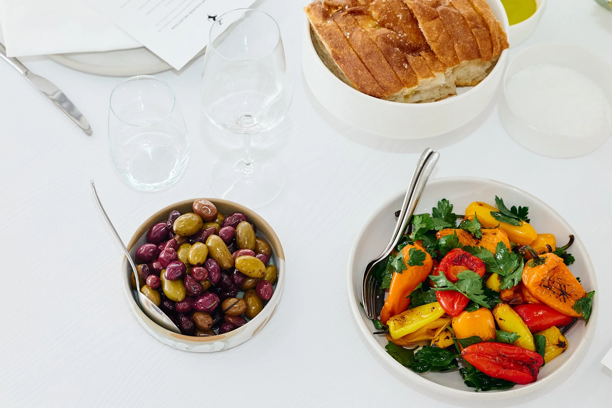 A white dining table with a colorful salad, a bowl of mixed olives, a bowl of sliced bread, an empty glass, a water glass, and a knife.